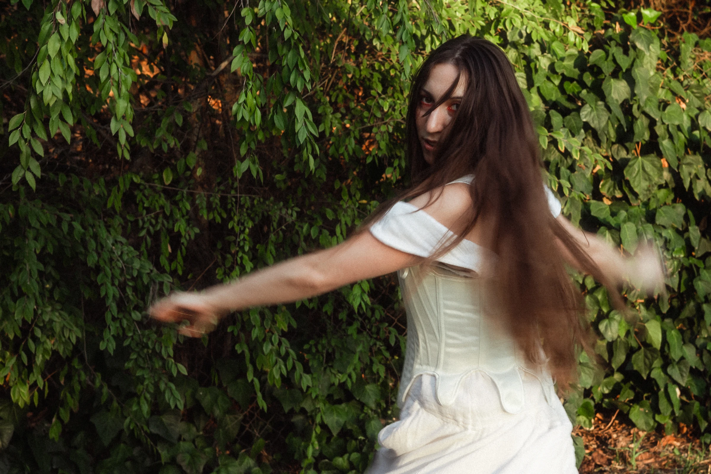 A woman with long hair twirls gracefully in a white dress against a lush, leafy backdrop. Her expression is serene, capturing a whimsical moment.