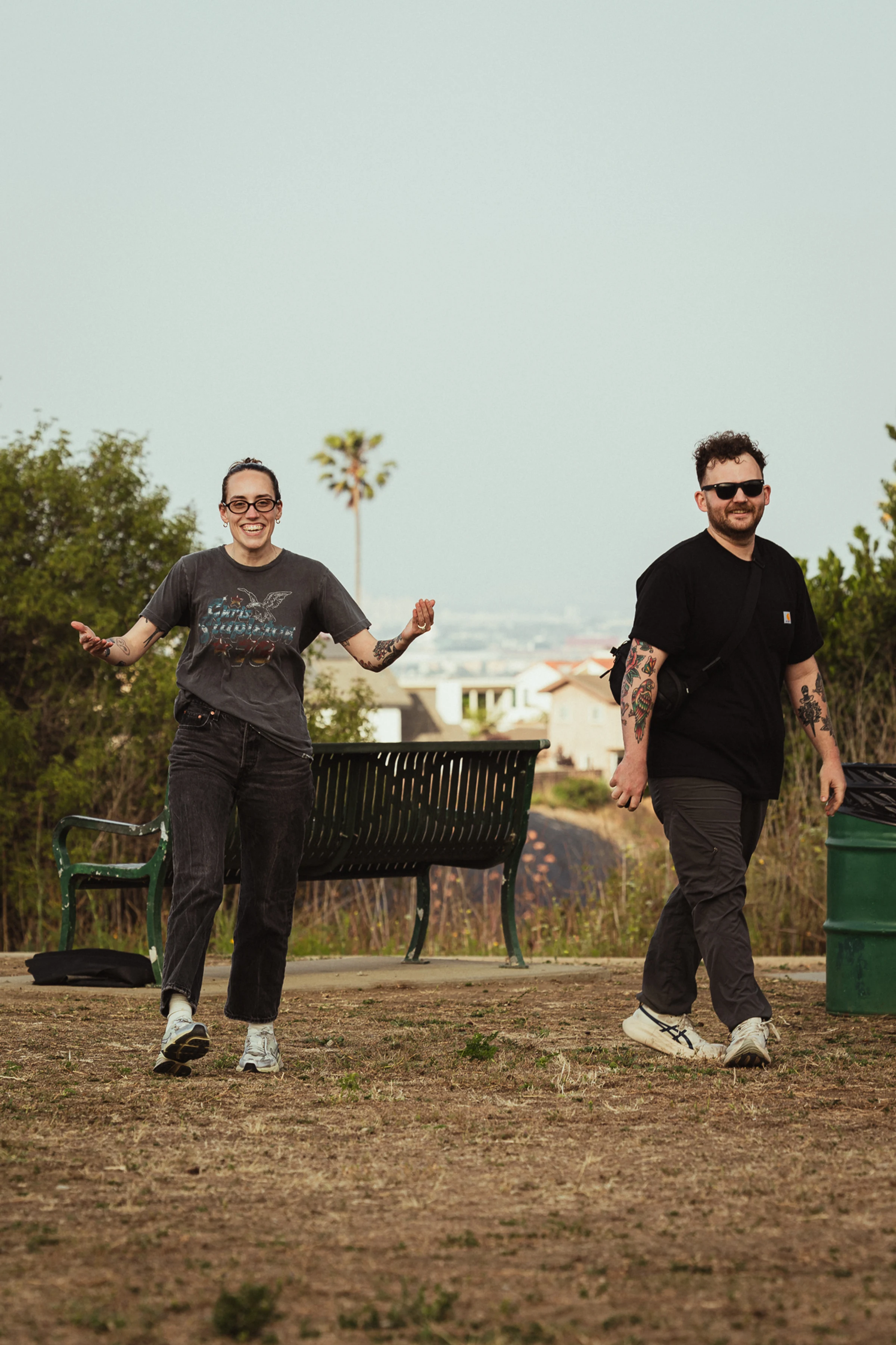 A smiling man and woman with tattoos walk happily on a sunny path, flanked by a green bench and trash can, with distant houses and a palm tree.