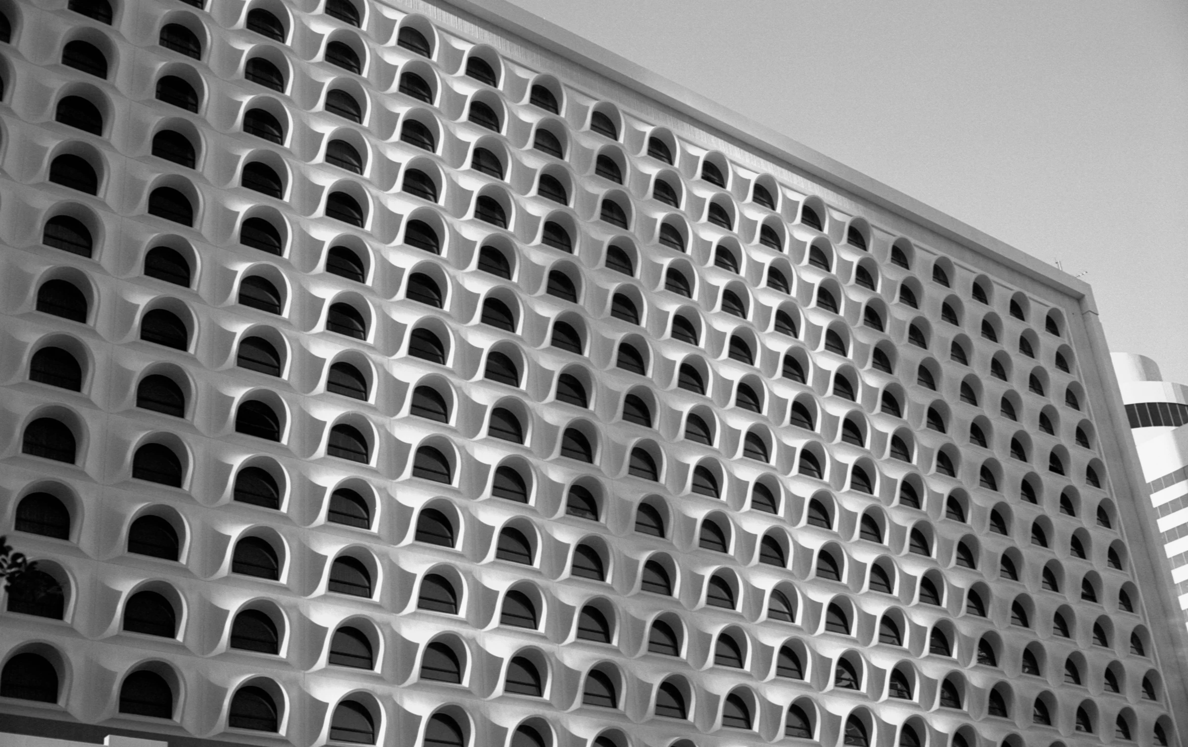 Modernist building facade with a grid of arched windows casting deep shadows. The monochrome image emphasizes the geometric, repetitive design.