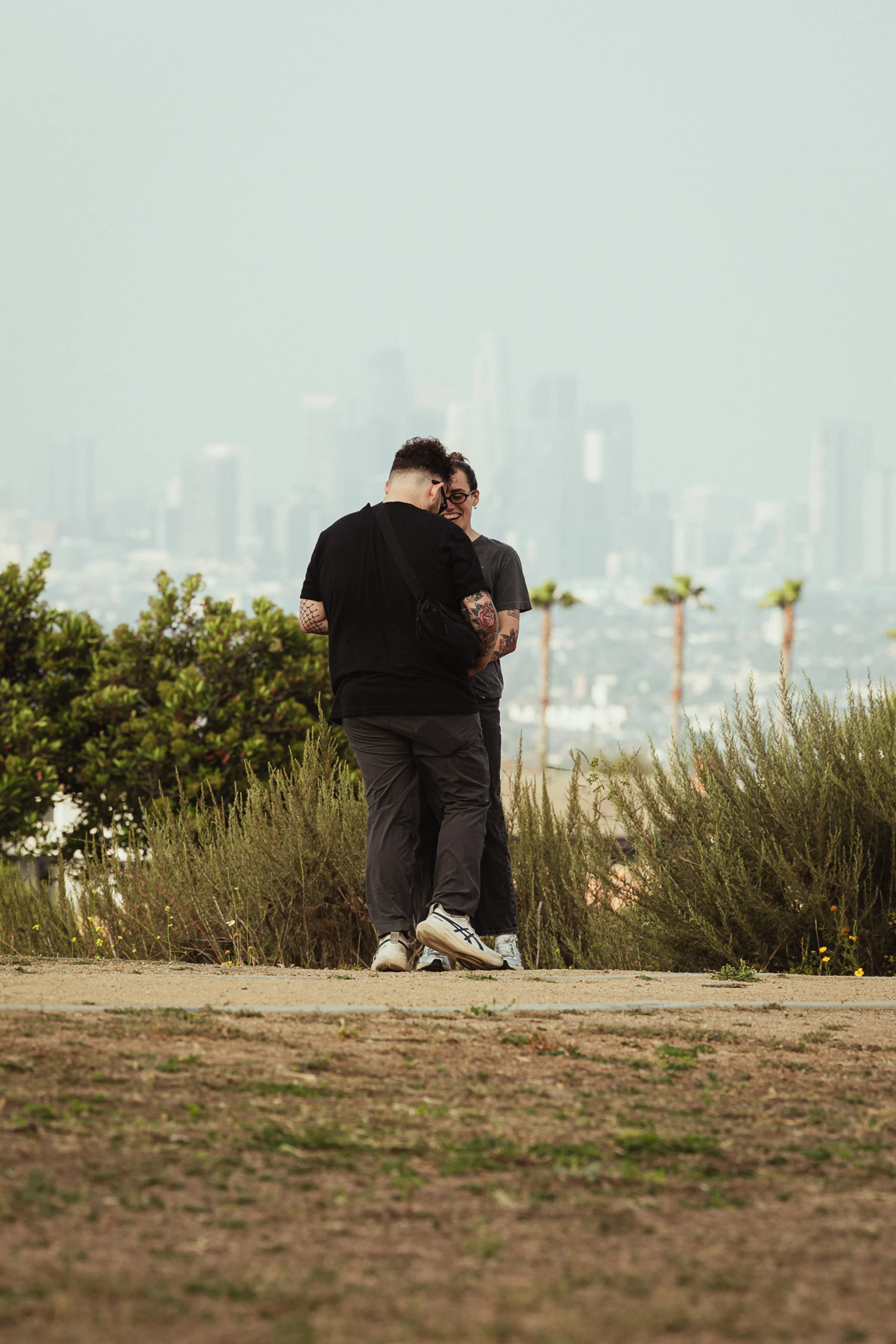 Two people stand facing each other on a grassy path, smiling and conversing. Palm trees and a hazy city skyline are visible in the background.