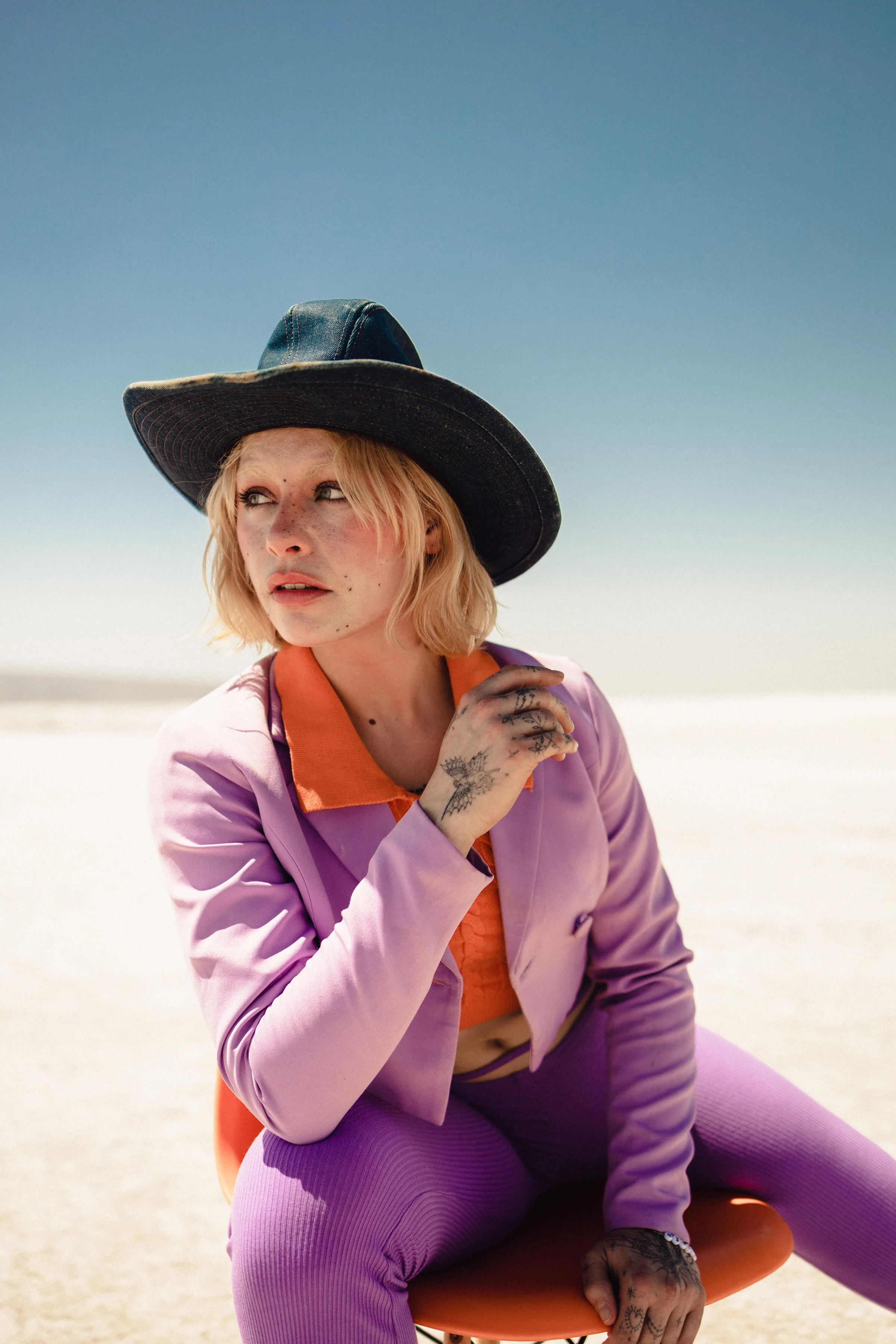 A person with blonde hair sits on an orange chair in a desert landscape, wearing a black cowboy hat and violet outfit, conveying a contemplative mood.