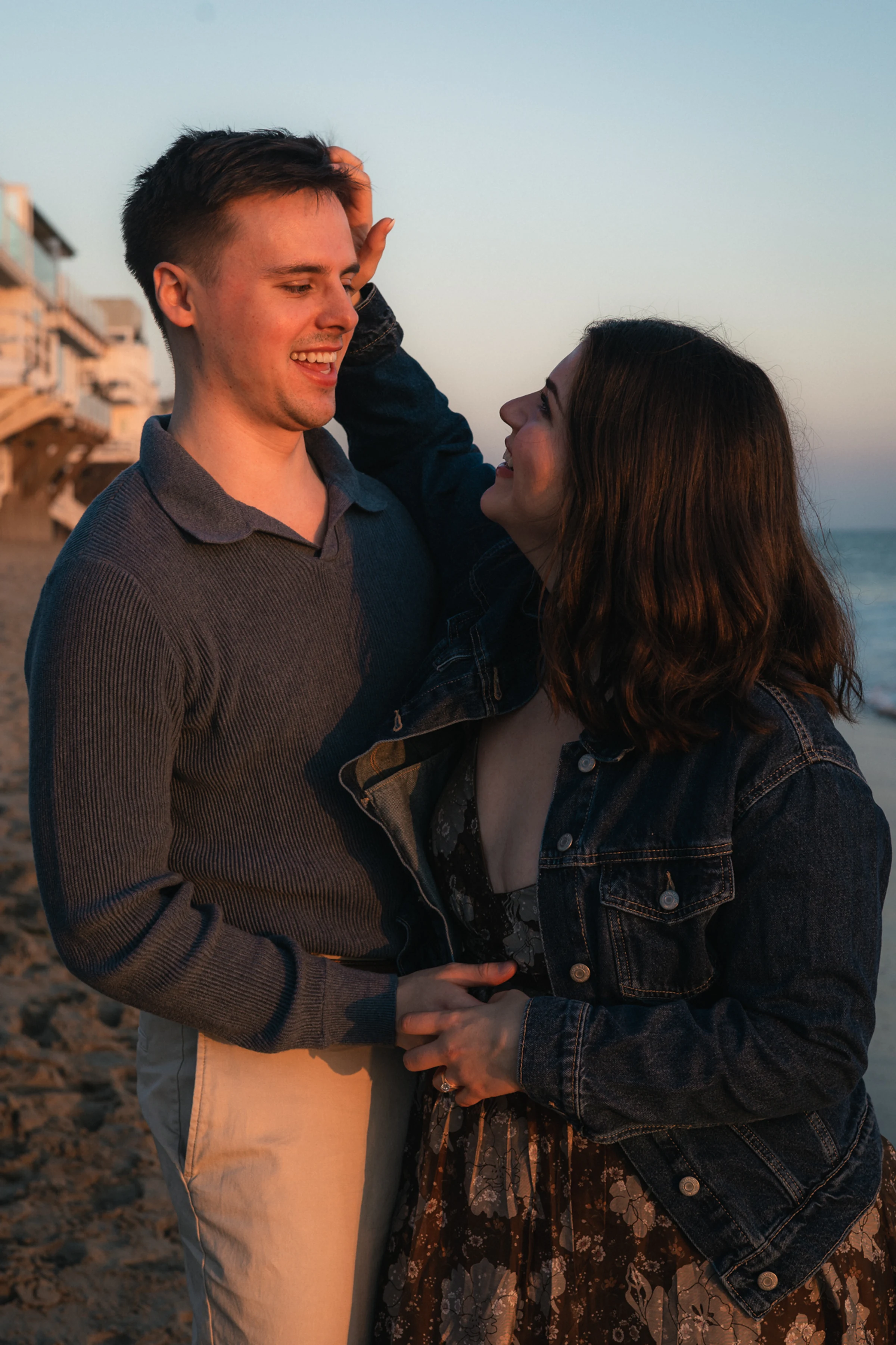 A couple laughs and gazes at each other on a sandy beach during sunset. The warm glow highlights their casual attire, creating a joyful, relaxed mood.