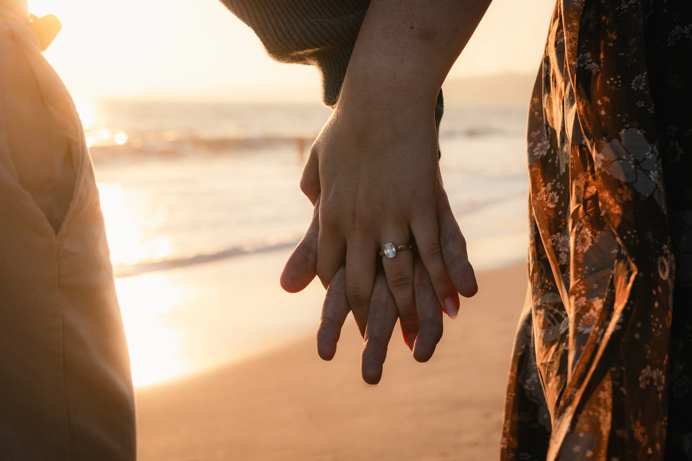 Close-up of a couple holding hands on a beach at sunset. A woman's hand shows an engagement ring, conveying a romantic and serene mood.