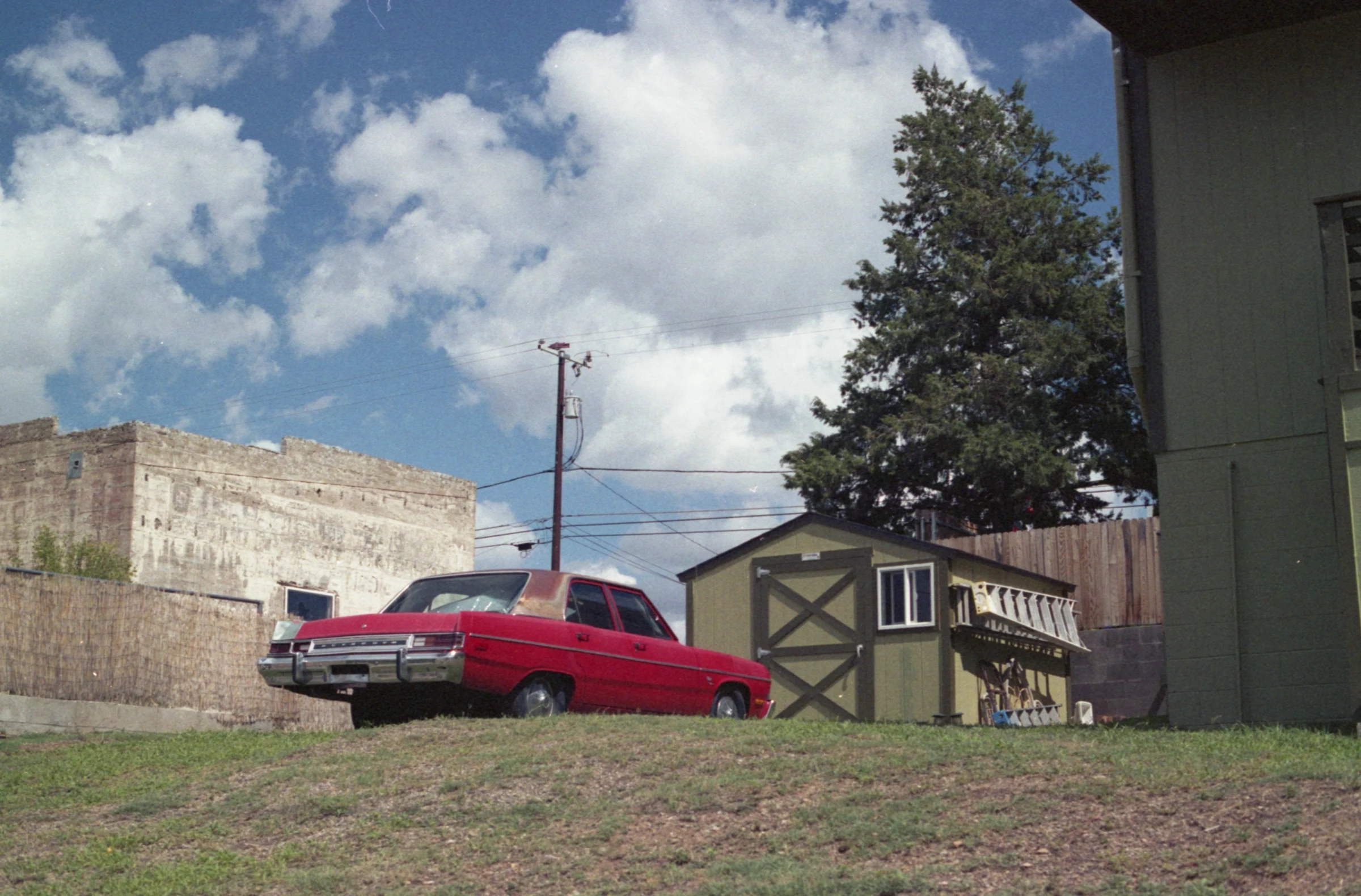 A red vintage car is parked on a grassy hill under a blue sky with fluffy clouds. Nearby are a wooden shed and an old brick building, conveying a nostalgic feel.