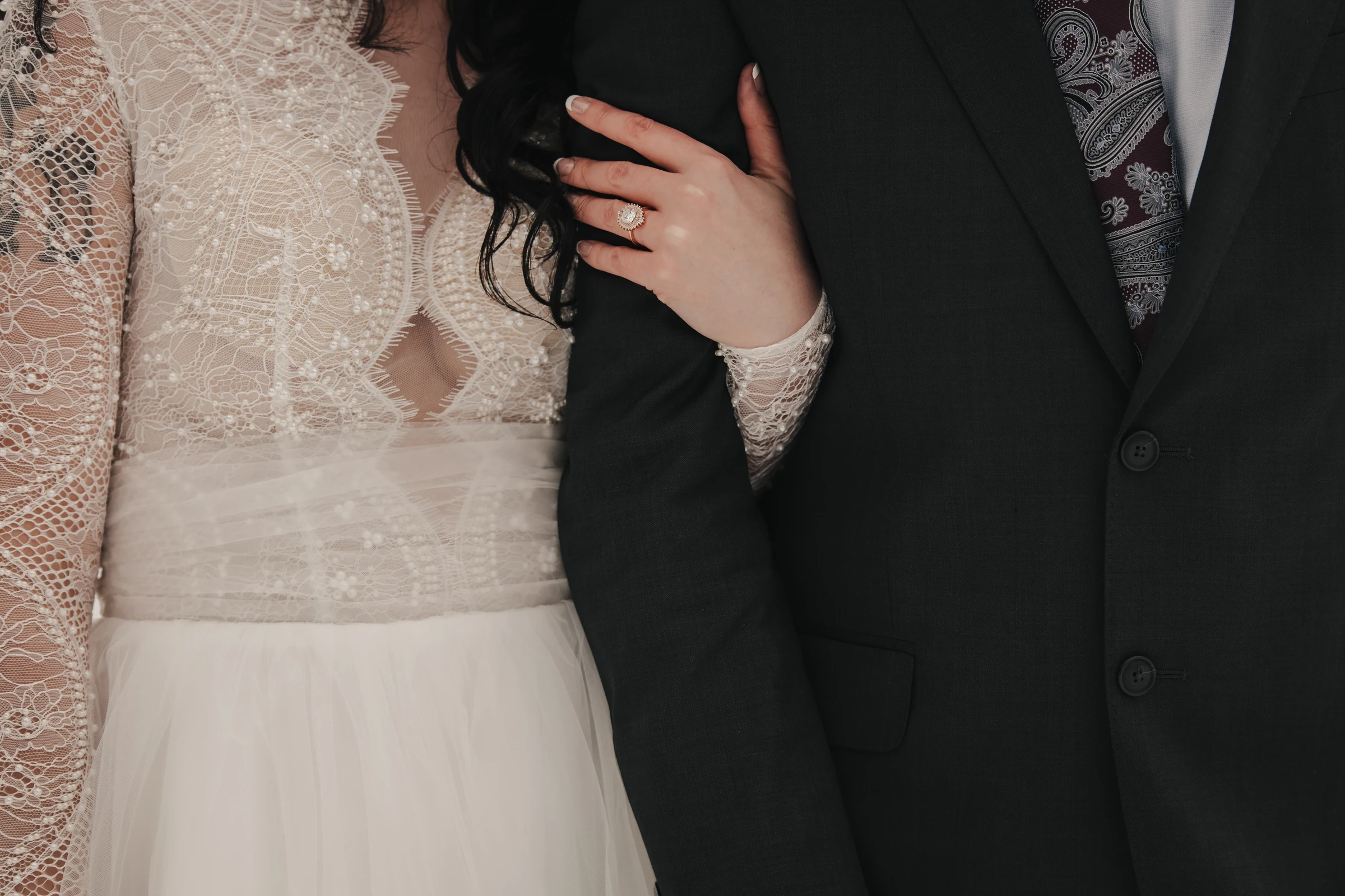 Bride in lace wedding dress holds groom's arm in a dark suit, showcasing her engagement ring. The image conveys elegance and intimacy.