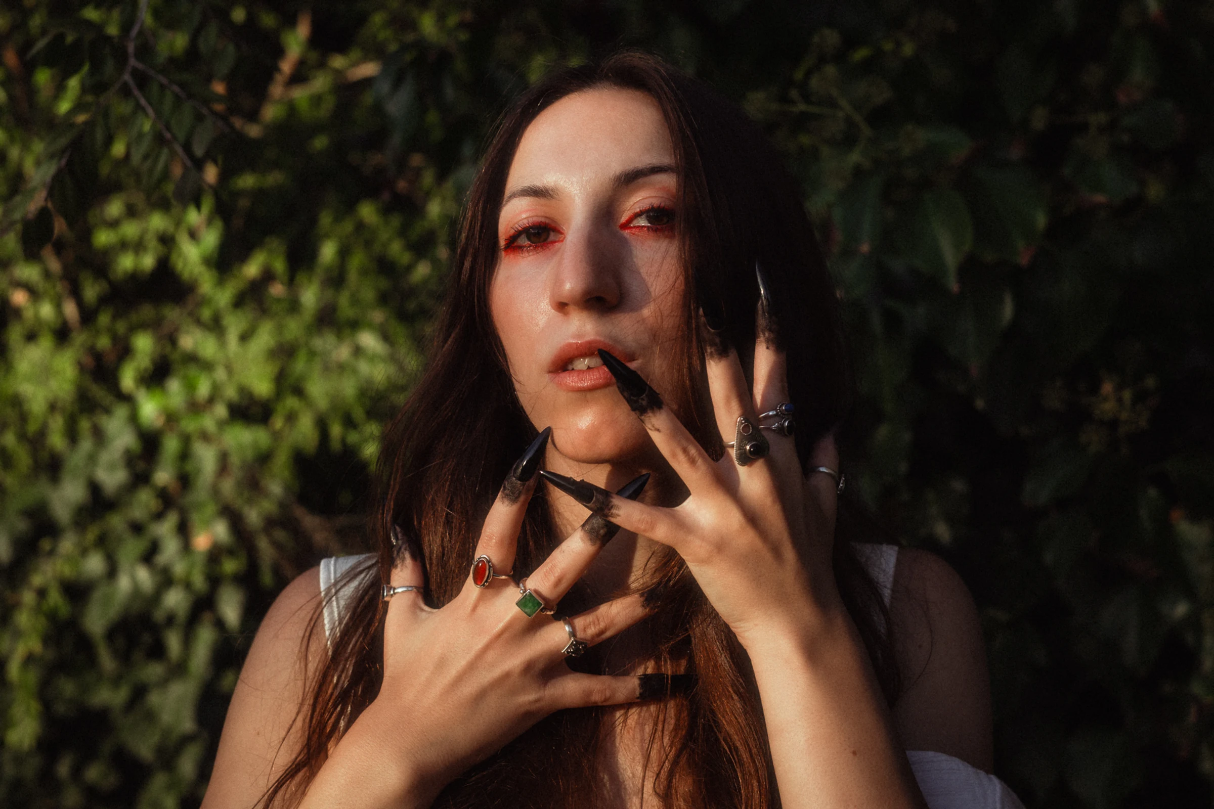 Woman with long dark nails and red eye makeup poses dramatically against a leafy background. Her expression is intense, and she wears multiple rings.