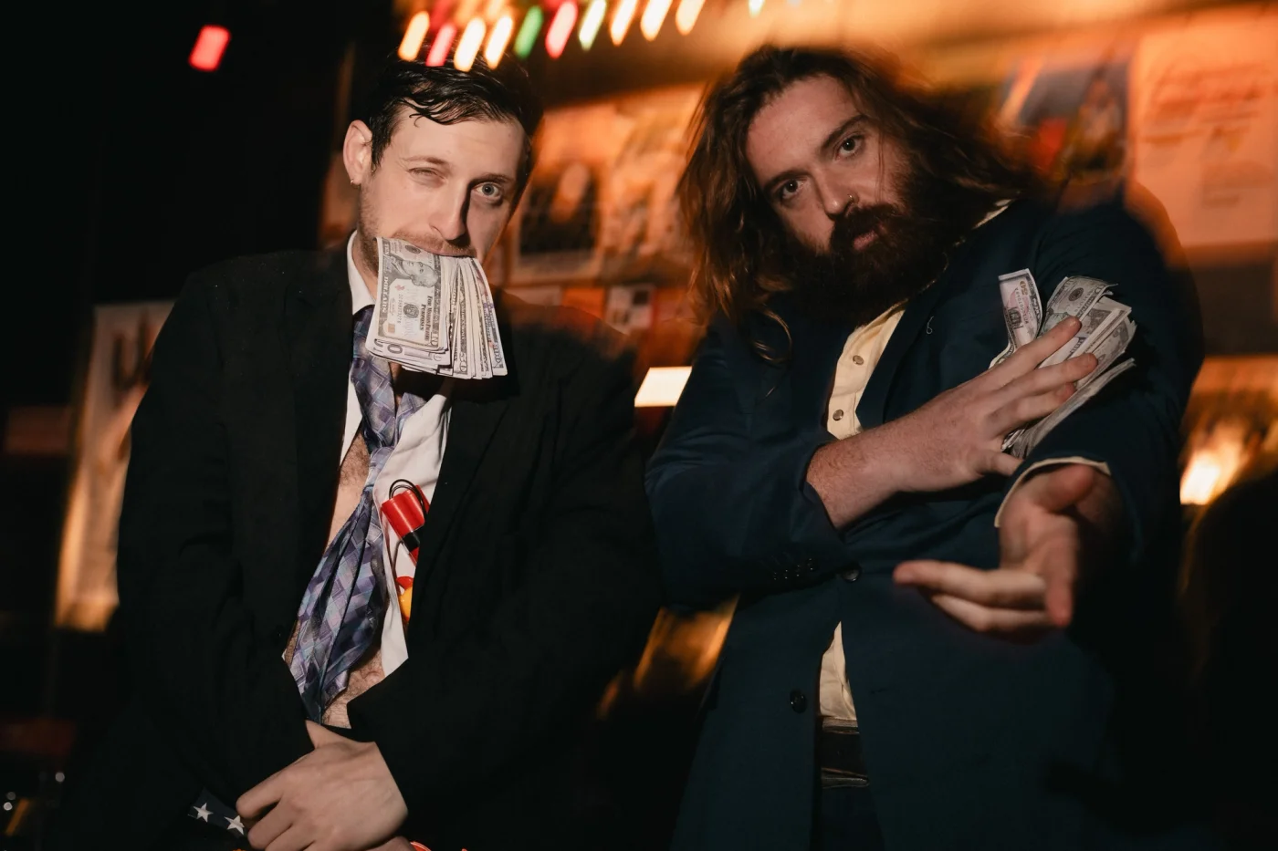 Two men in suits pose humorously, holding dollar bills in a dimly lit room with colorful lights. One has money in his mouth, both with playful expressions.