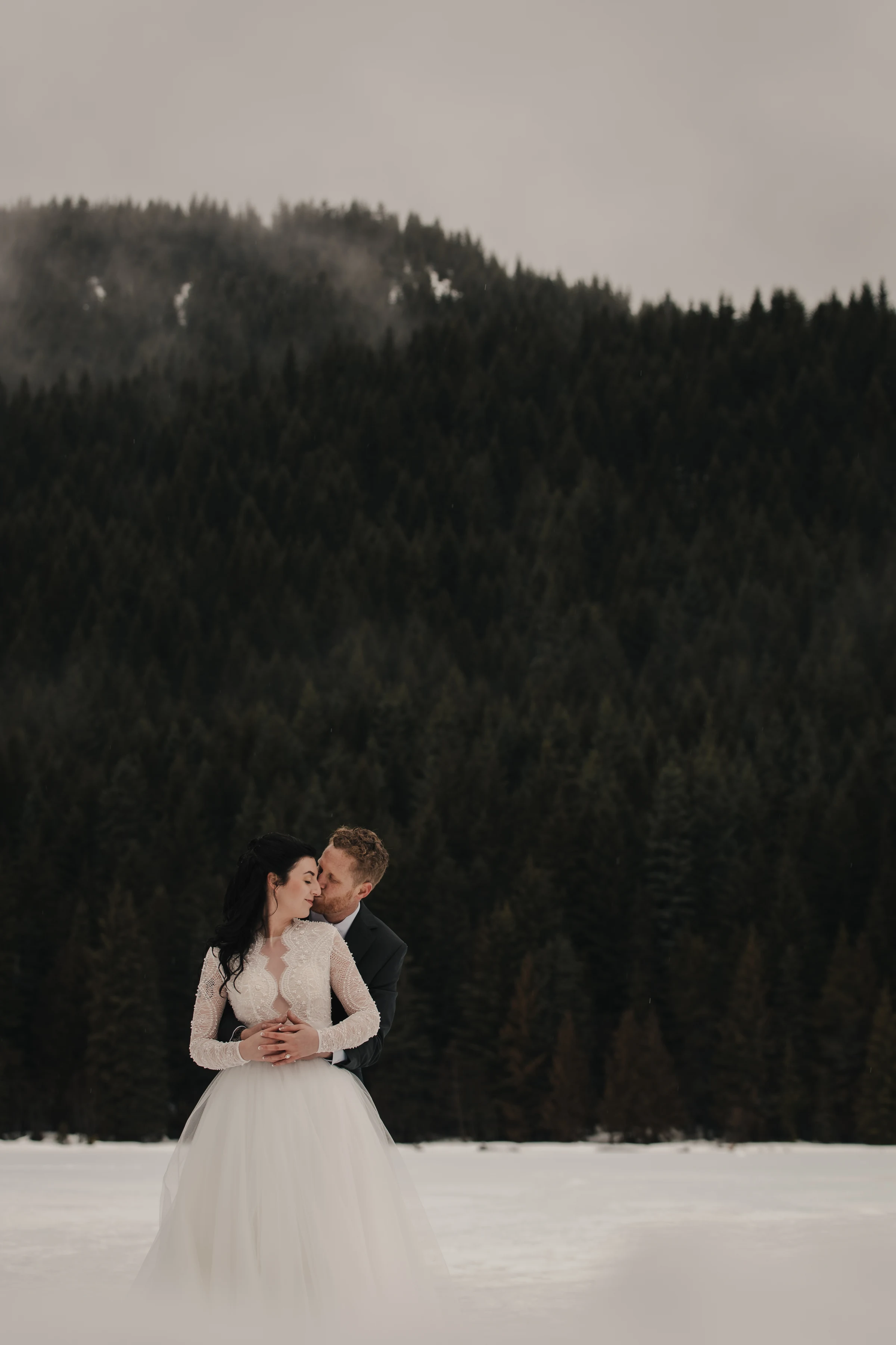 A couple embraces in a snowy landscape, the bride in a white dress smiles, while the groom in a dark suit gazes lovingly at her. A dense, misty forest serves as the backdrop.