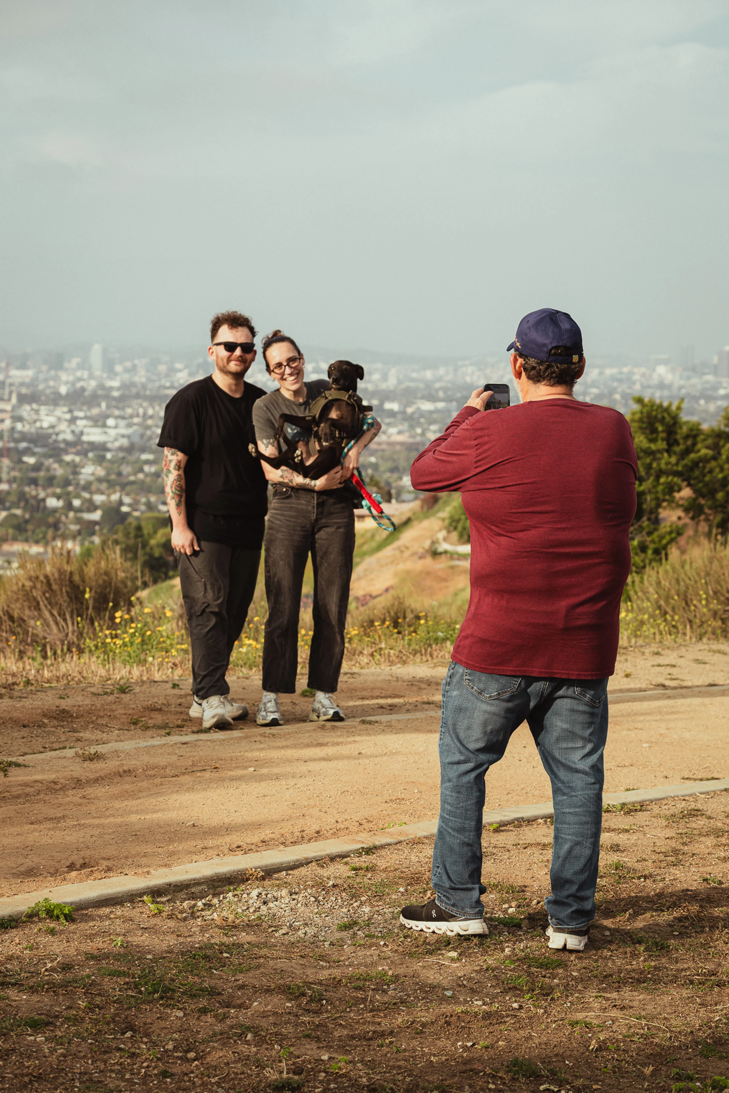 Two people with a dog pose for a photo on a scenic hilltop trail. Another person stands in the foreground, capturing the moment on a phone.