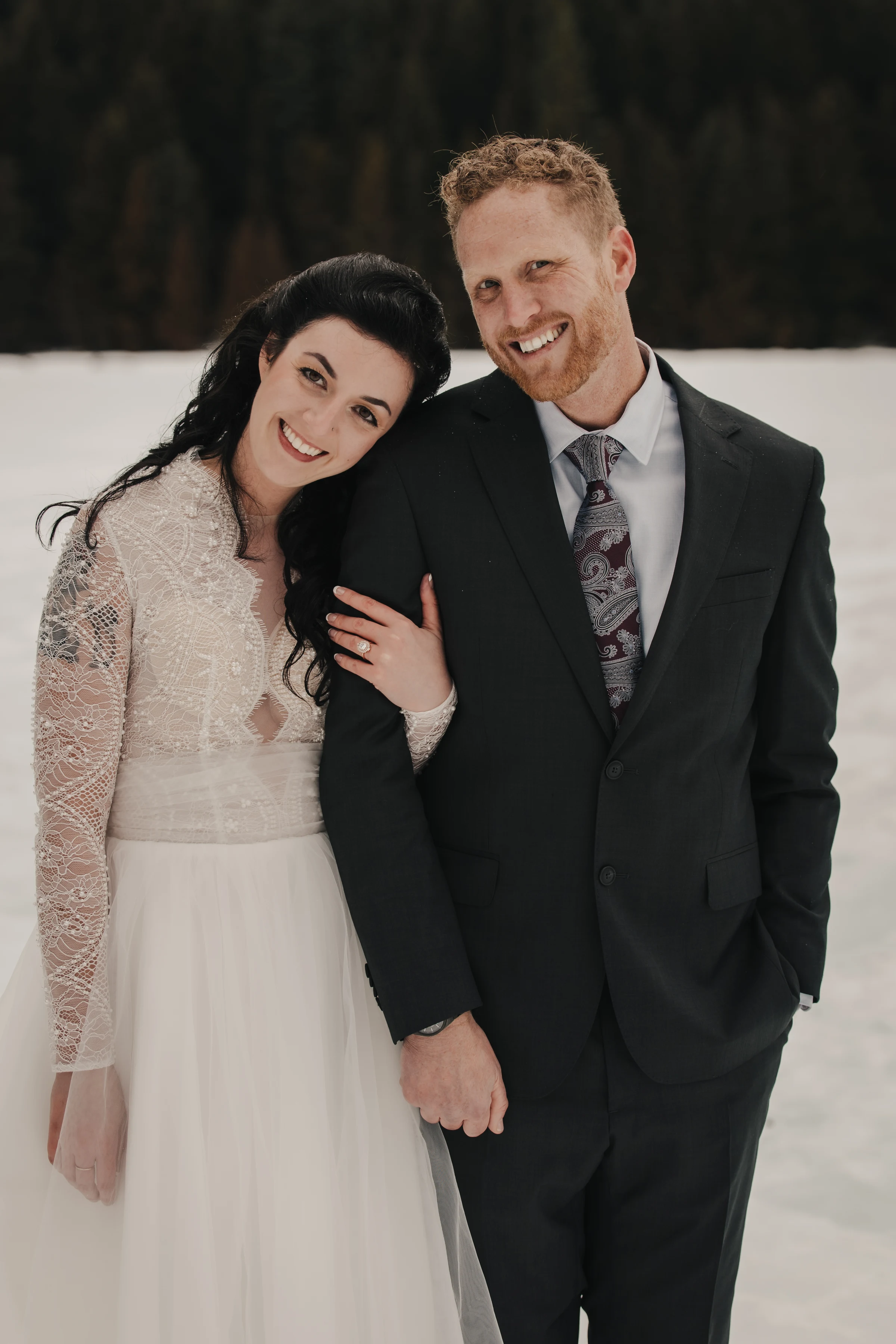 A bride in a lace gown and groom in a dark suit smiling warmly, standing close on a snowy landscape with a forested background. The mood is joyful and romantic.