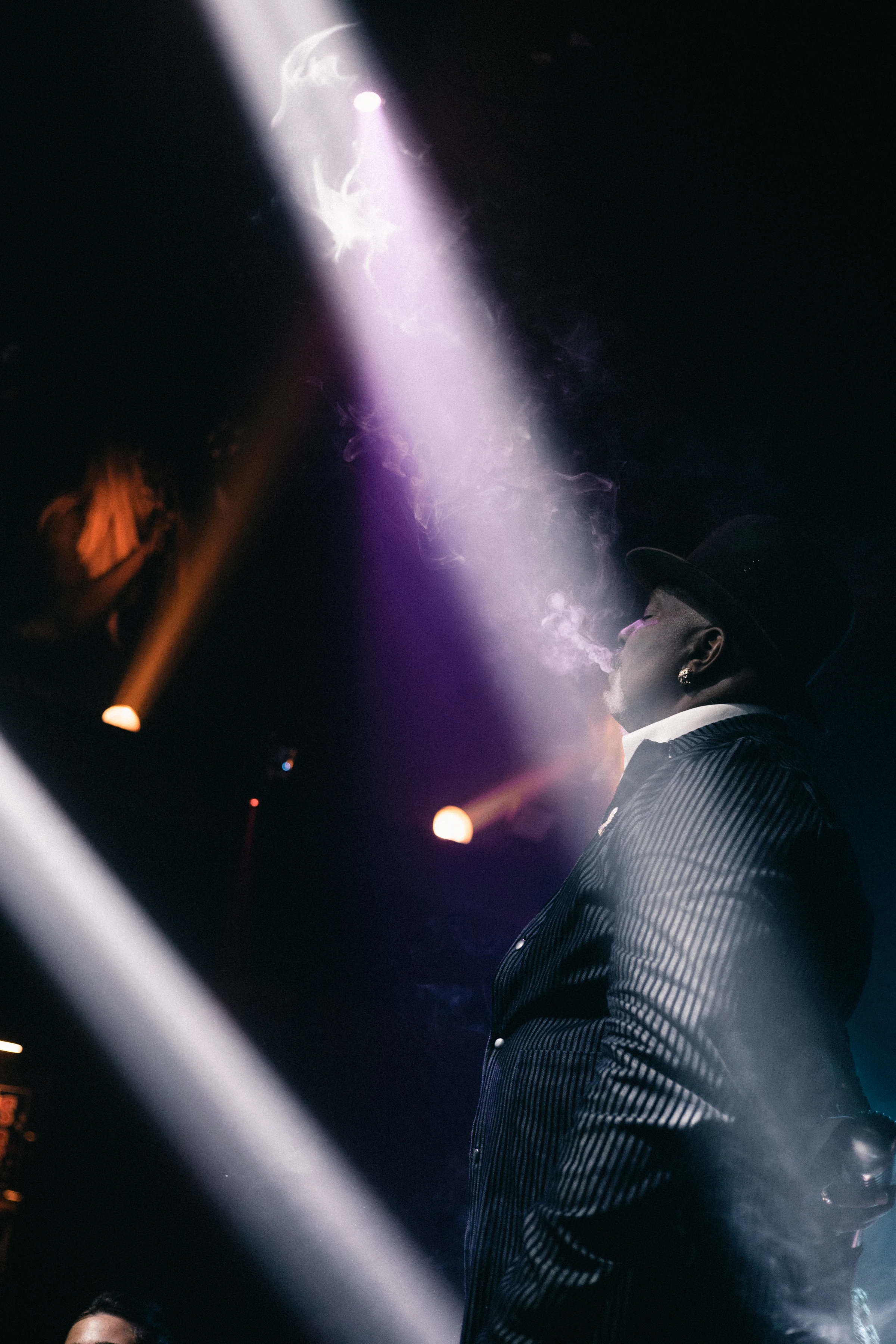 A person in a pinstripe suit and hat exhales smoke under dramatic stage lighting, with purple and orange beams creating a moody, atmospheric effect.