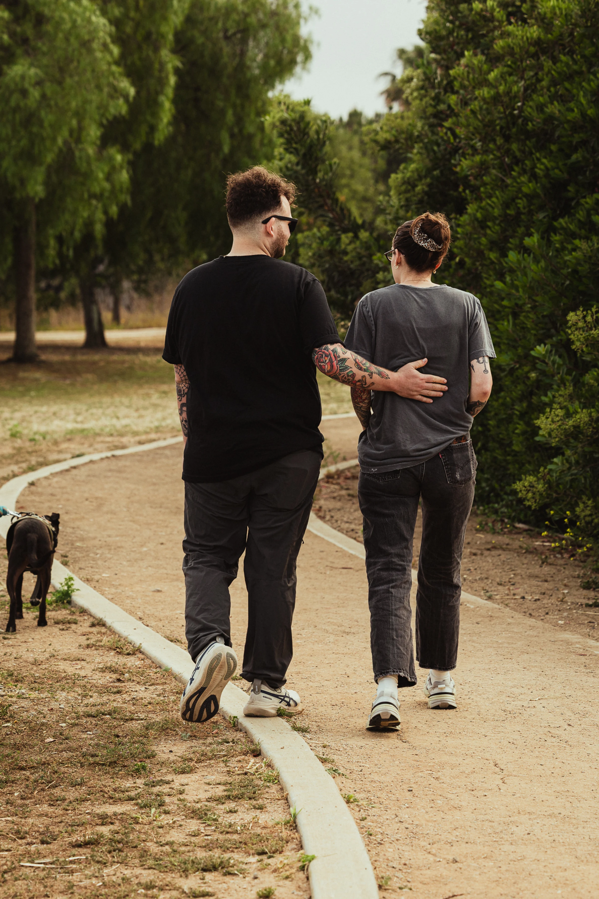 Two people walk on a tree-lined path, one arm around the other. A black dog walks ahead. The scene conveys companionship and tranquility.