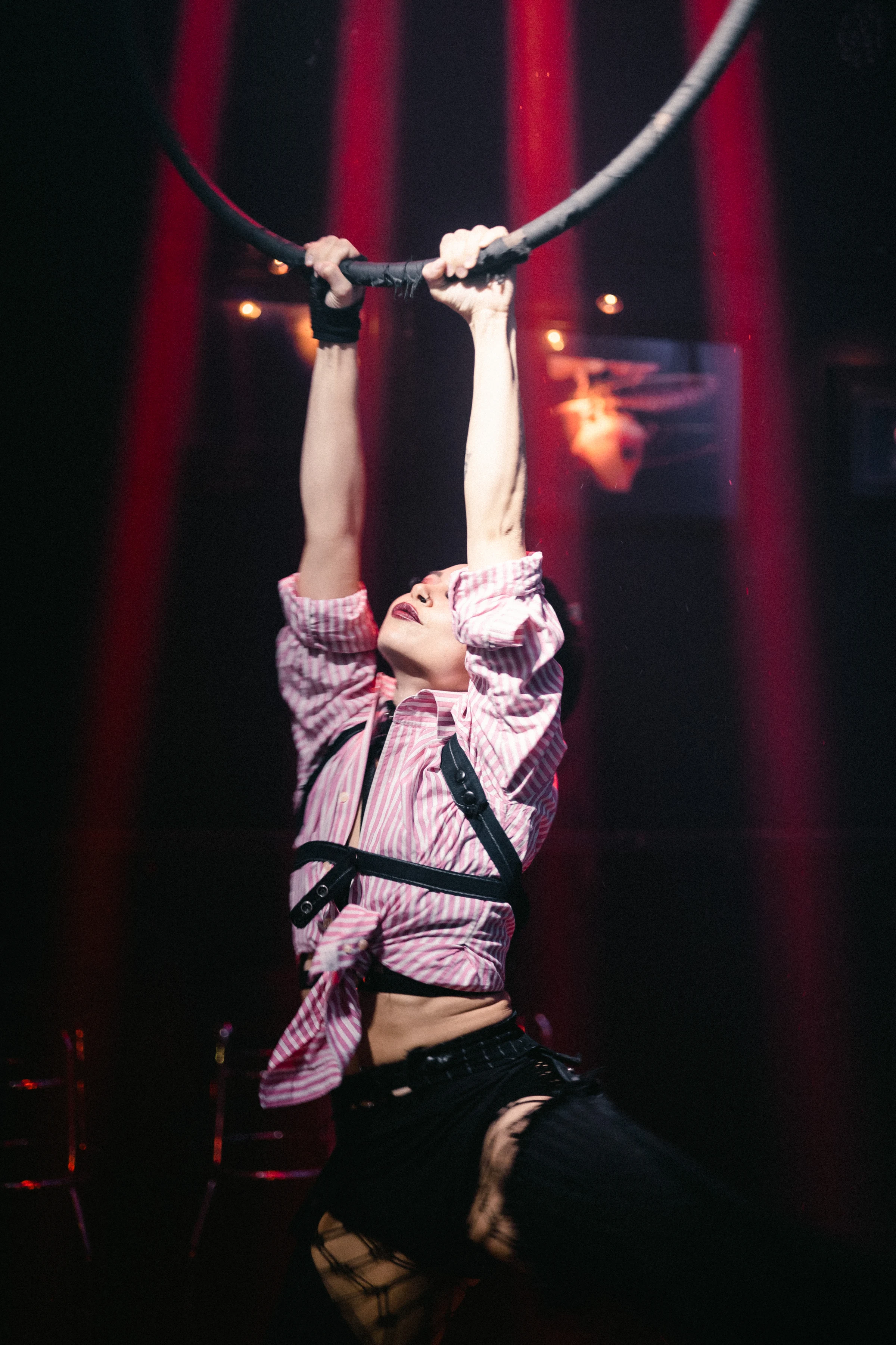 A performer in a striped shirt and harness grips a ring, suspended in mid-air, under dramatic red and pink stage lights, exuding strength and focus.