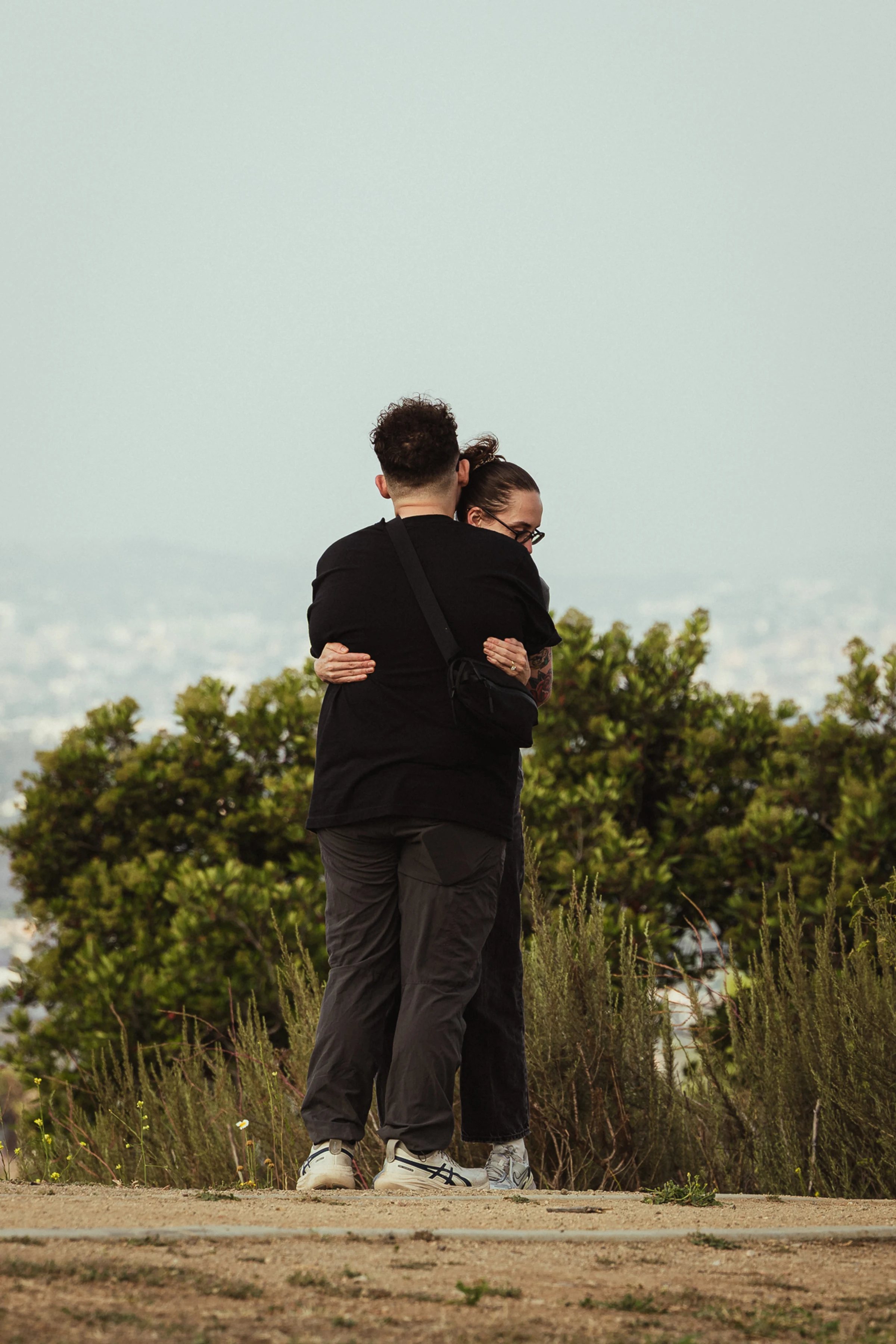 Two people in black clothing share a warm embrace outdoors, surrounded by greenery. The background features a blurred cityscape under a clear sky.