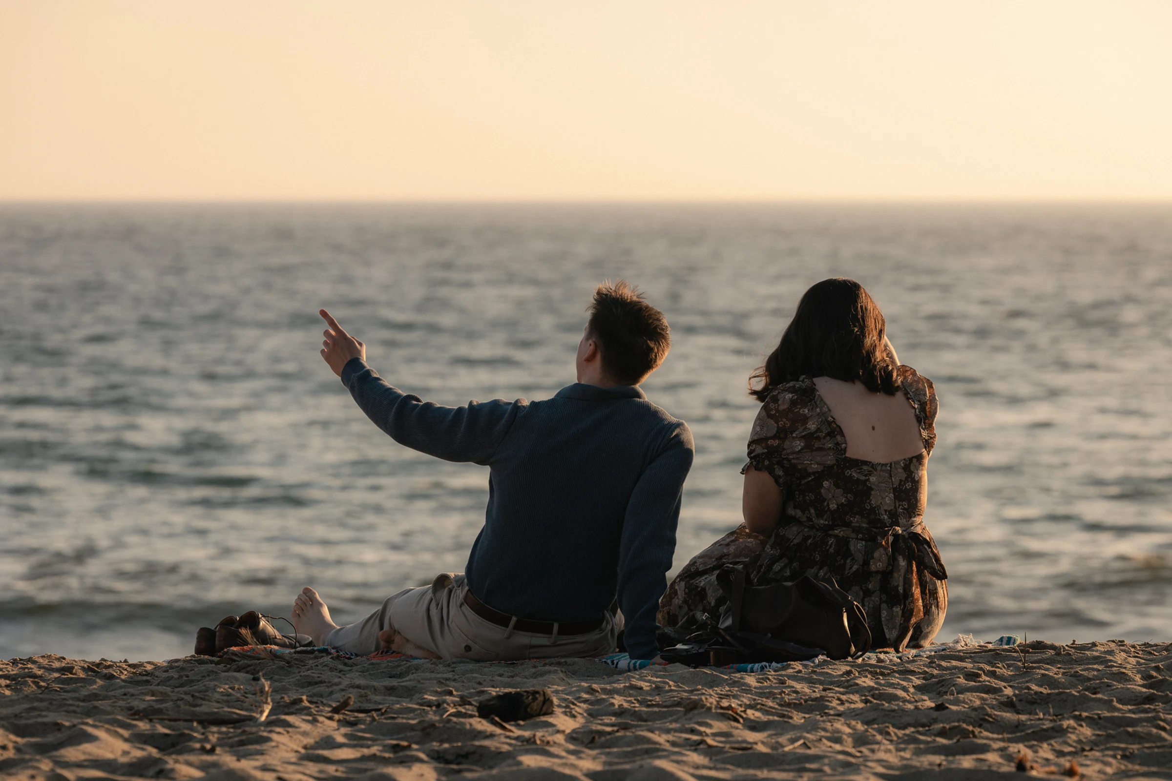 A couple sits on a sandy beach at sunset, facing the calm ocean. The man points towards the horizon. The scene is peaceful and reflective.