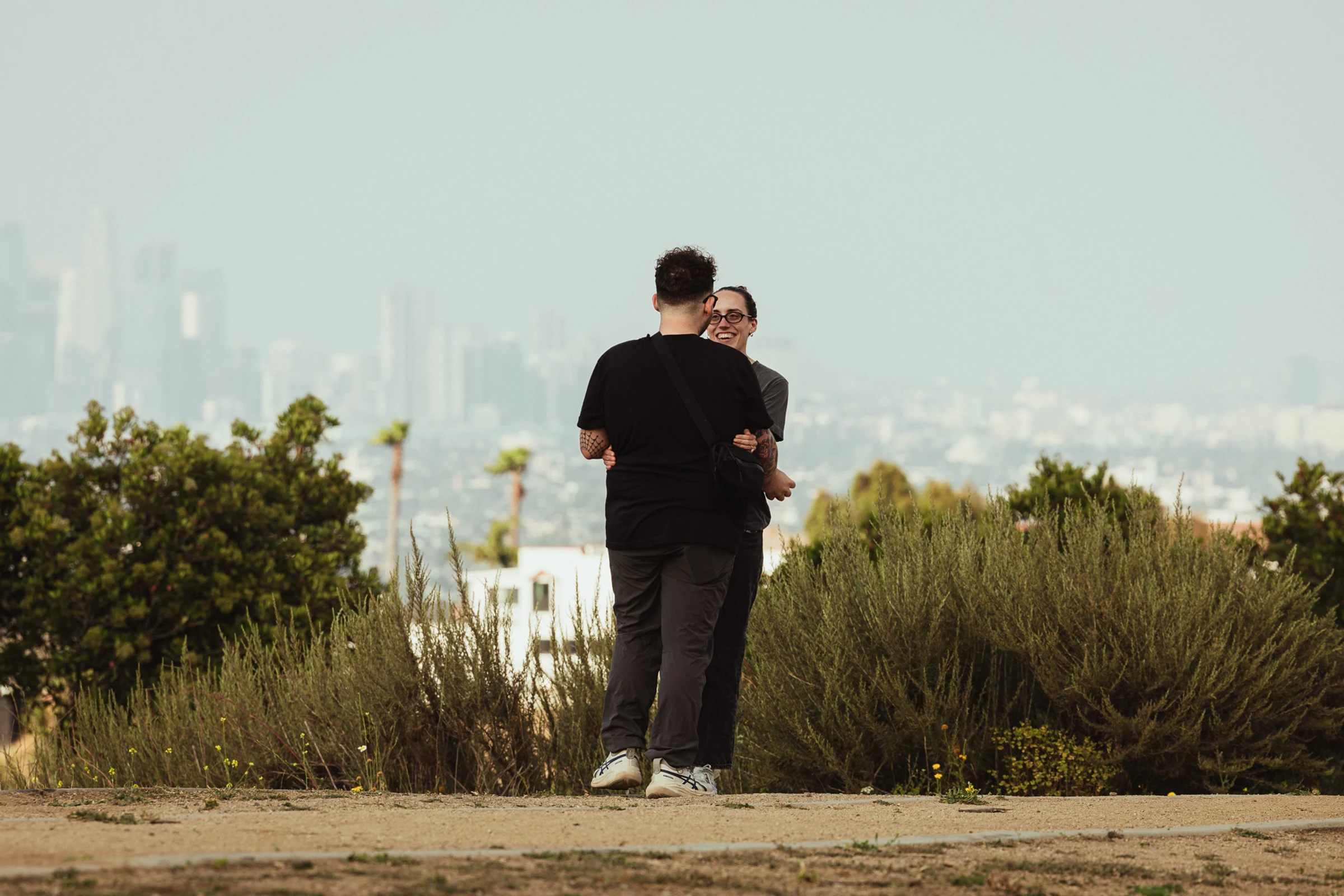 Two people stand closely on a dirt path, smiling warmly at each other. Behind them, green shrubs and a blurred cityscape are visible under a clear sky.