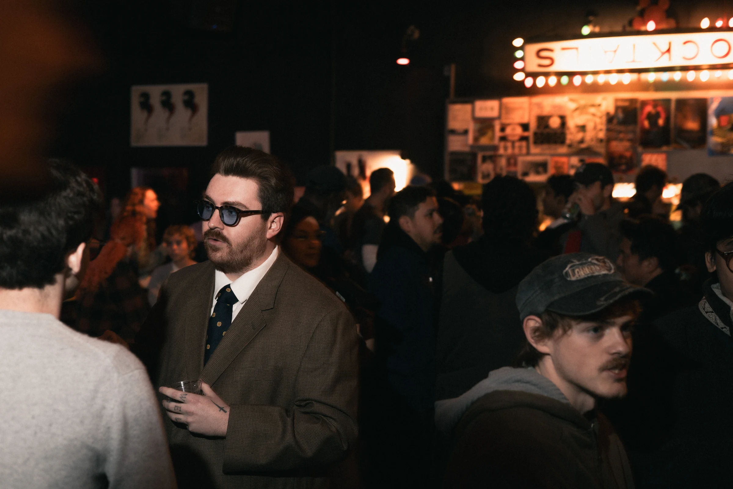 A man in a suit with sunglasses holds a drink in a dimly lit, crowded bar. Neon sign and posters adorn the background, conveying a lively atmosphere.
