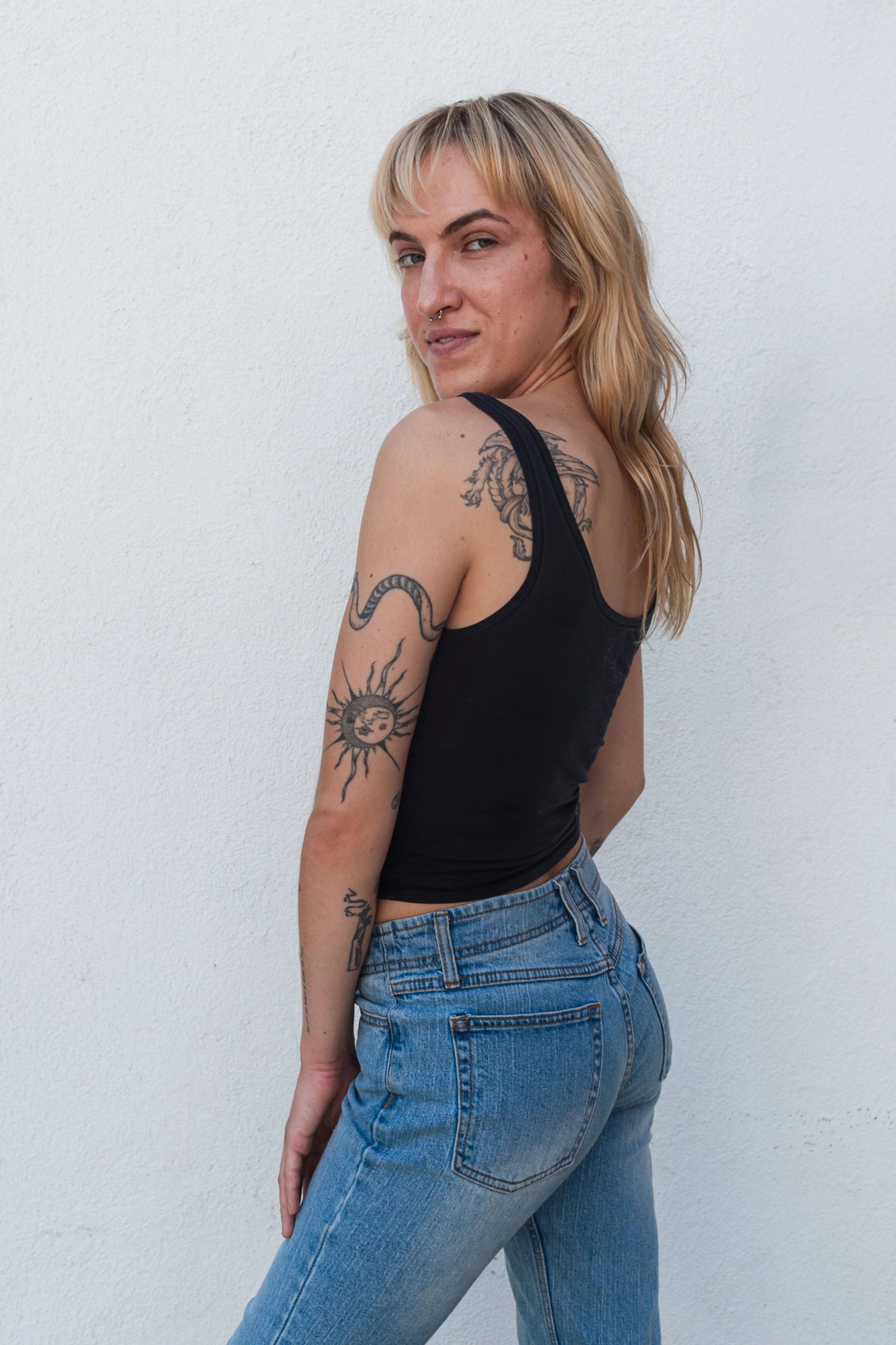 A woman with long blonde hair and tattoos poses confidently against a white wall, wearing a black tank top and blue jeans, exuding a casual vibe.