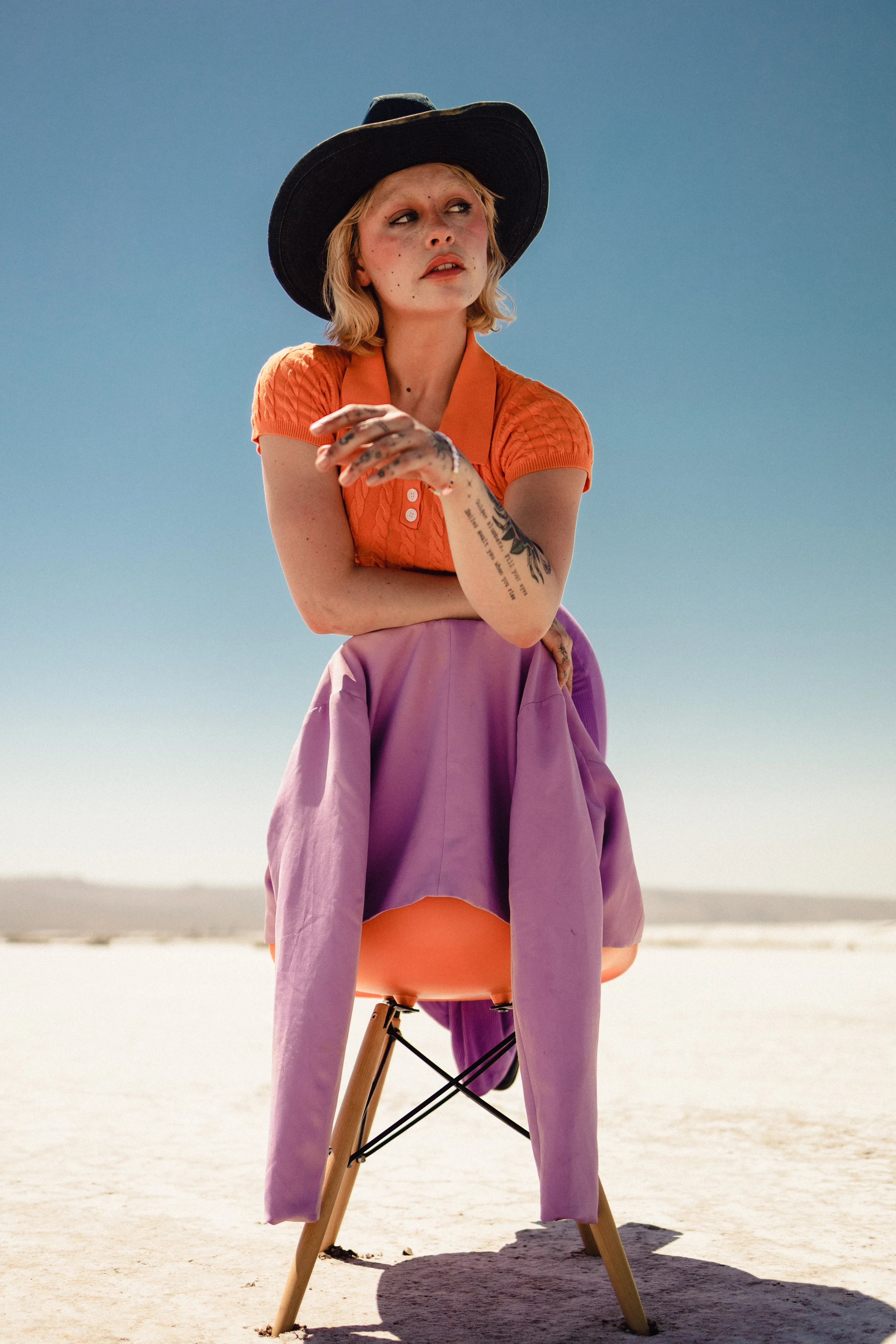 A person sits outdoors on a chair in a desert, wearing a black hat, orange top, and purple skirt. The sunny scene evokes a stylish, contemplative mood.
