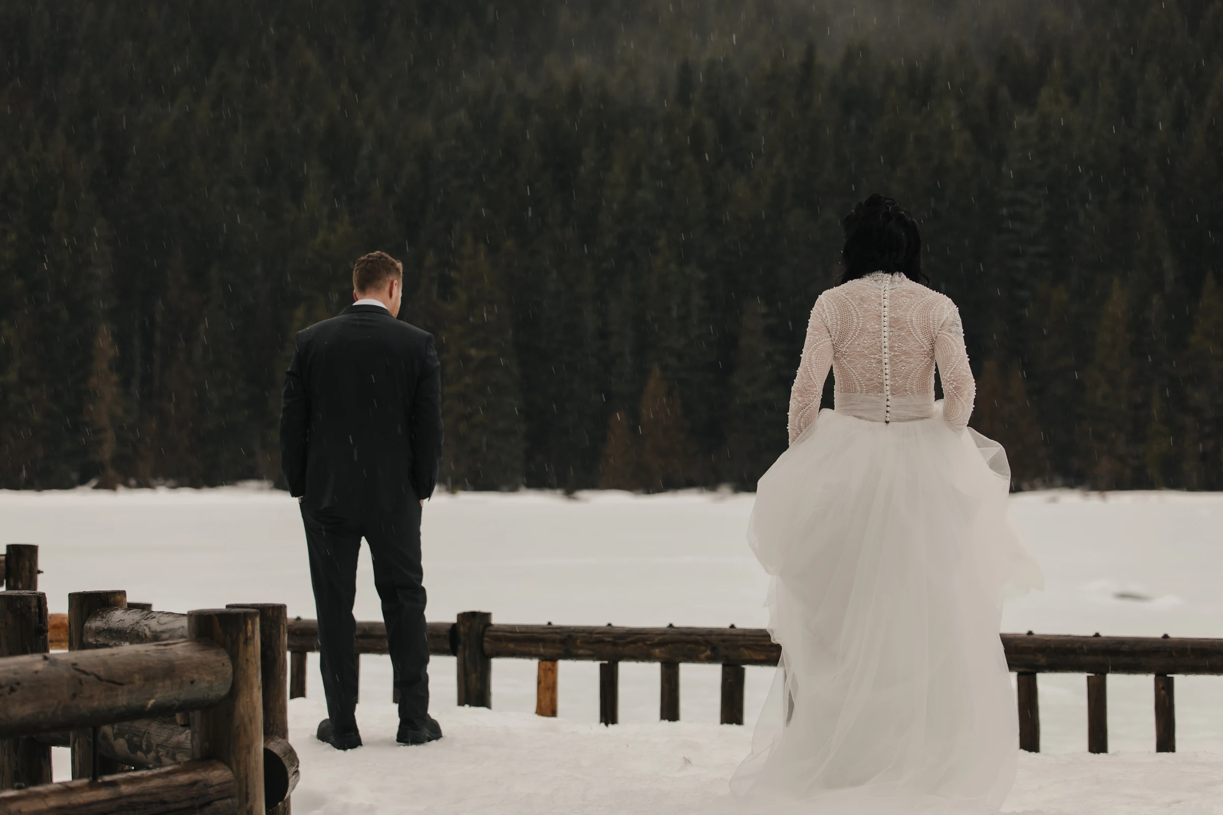 Bride in a lace gown and man in a suit stand on snowy ground facing a serene, forested landscape, evoking a mood of quiet reflection and romance.