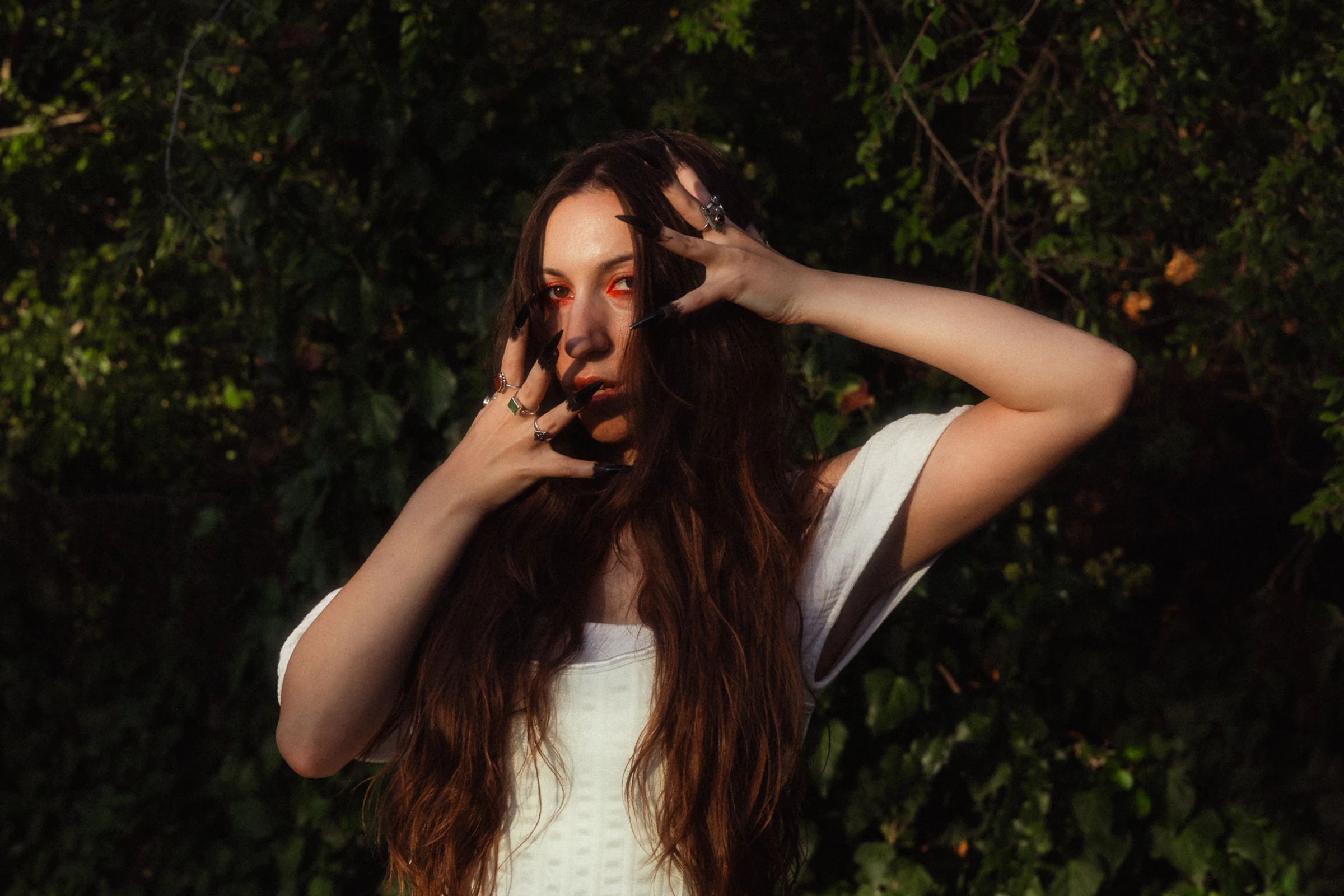A woman with long brown hair and red eye makeup poses outdoors, surrounded by lush green foliage. She wears a white top and holds her hands near her face, conveying a mysterious and dramatic mood.