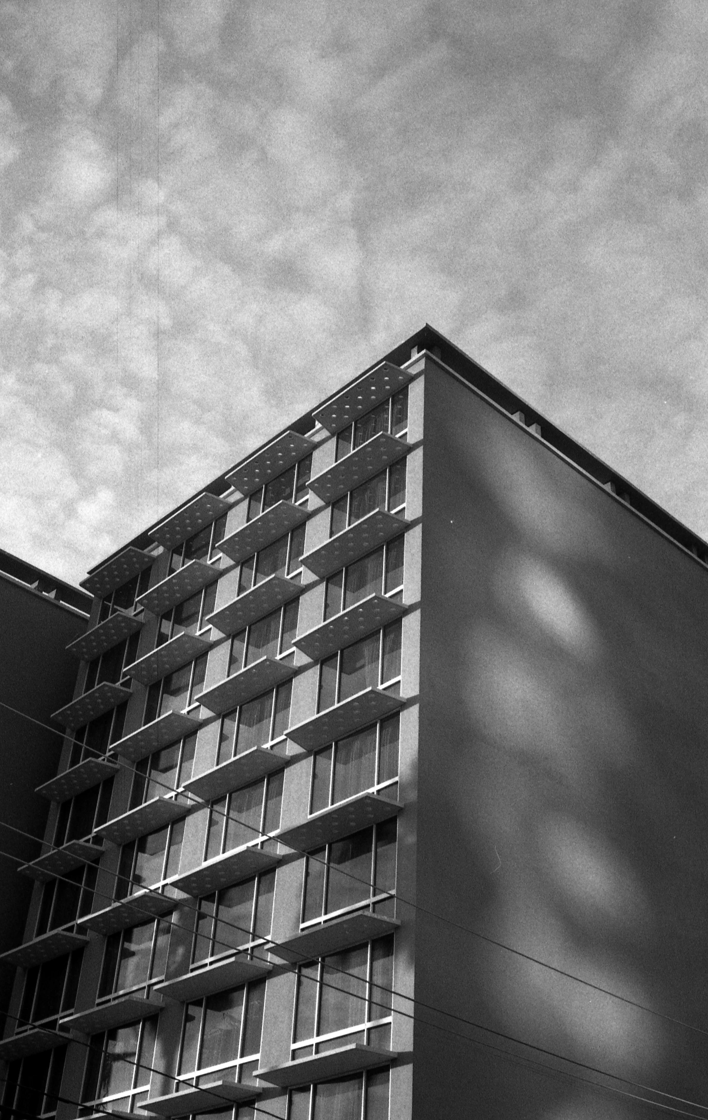 Black and white photo of a modern building with a grid of windows and awnings. Shadows on the facade create a geometric pattern, set against a textured cloudy sky.