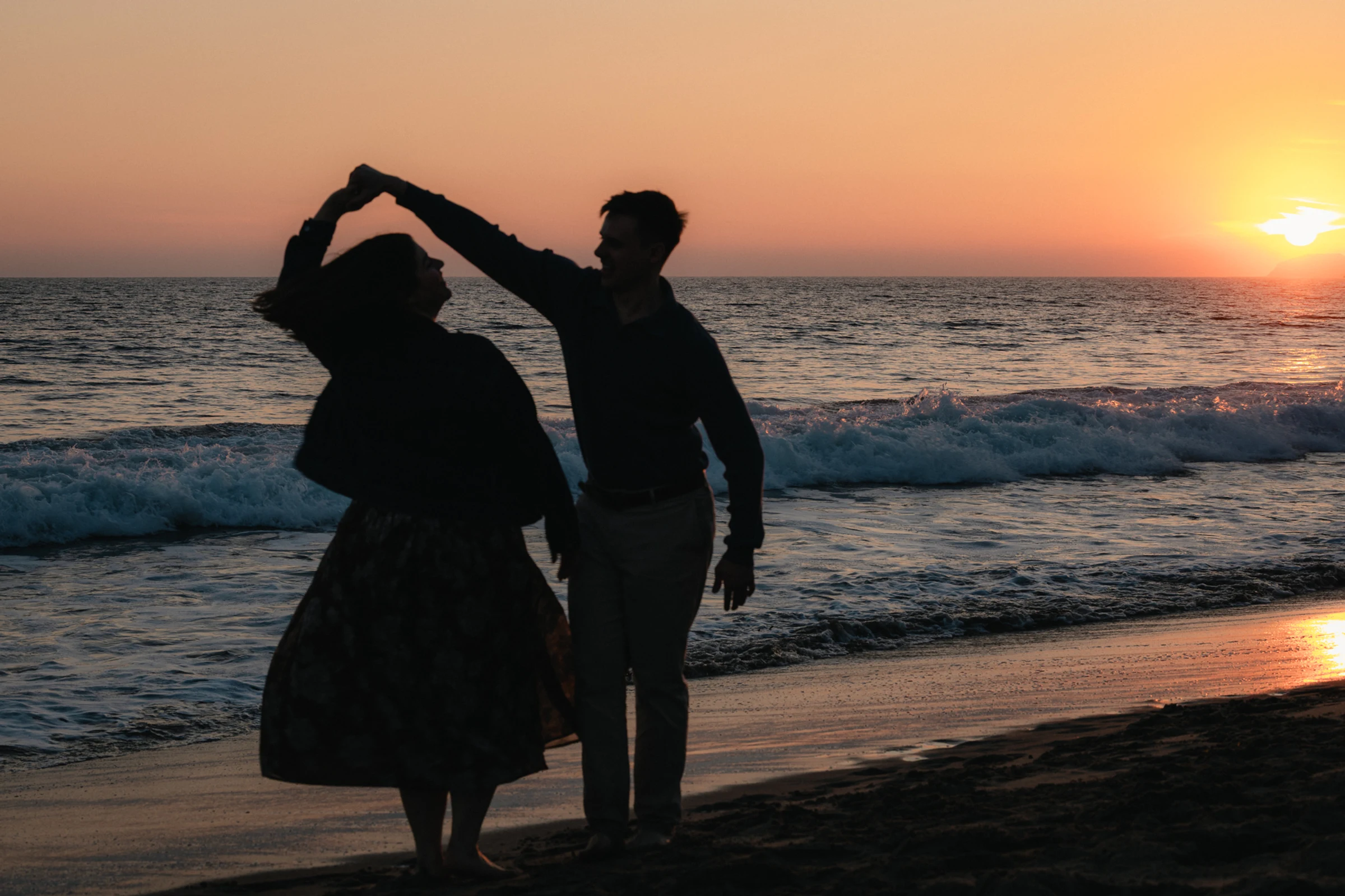 A couple dances playfully on a beach at sunset, silhouetted against the colorful sky. Waves gently crash onto the shore, creating a romantic atmosphere.