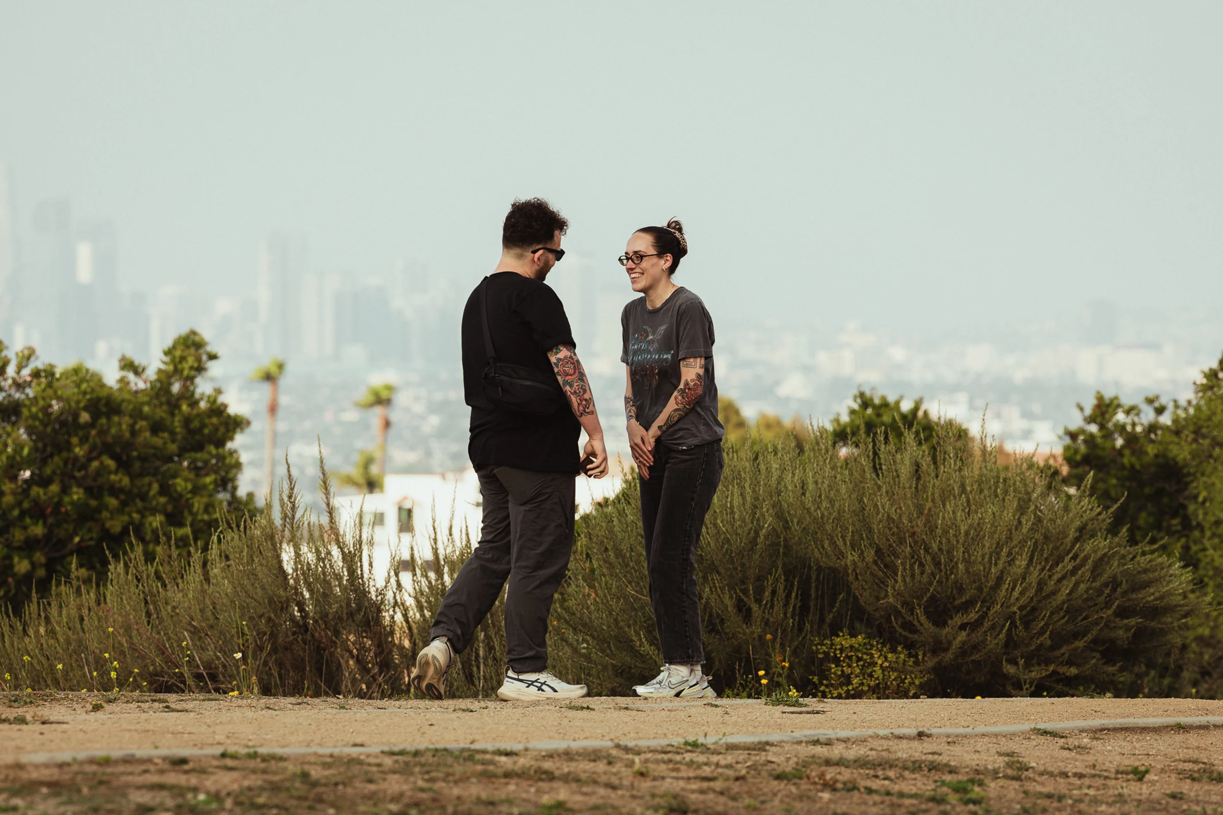 A man and woman stand on a dirt path in a park, smiling at each other. They're surrounded by green shrubs, with a city skyline in the hazy distance.