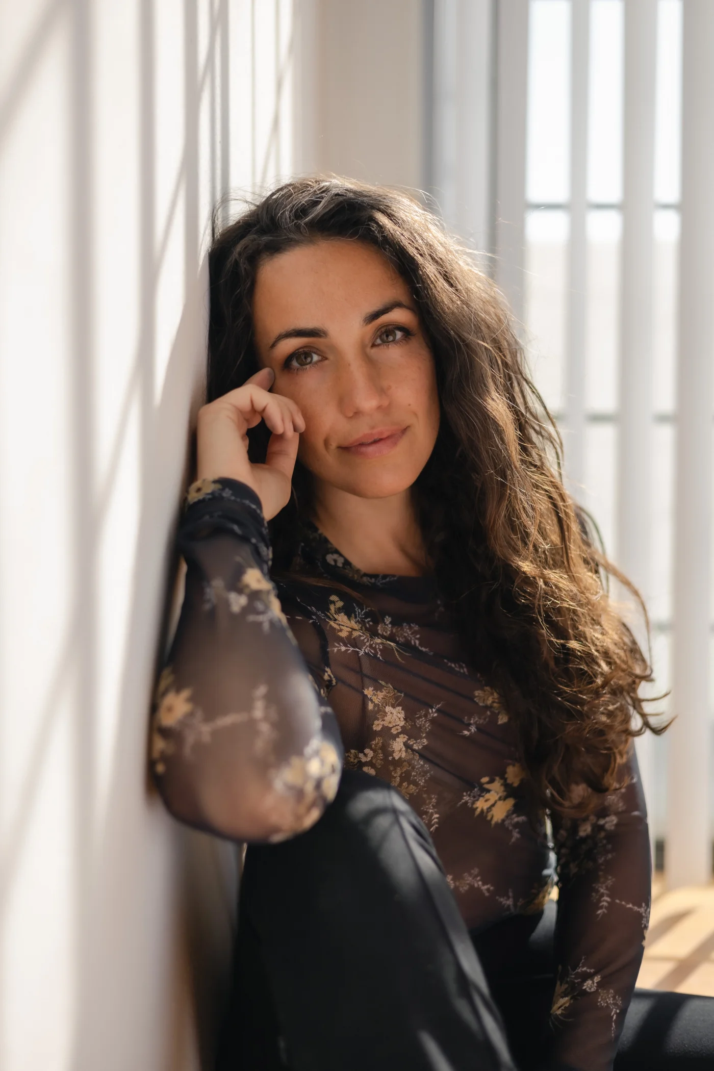 A woman with long wavy hair leans against a sunlit wall. She's wearing a sheer floral top, exuding a calm and contemplative mood.