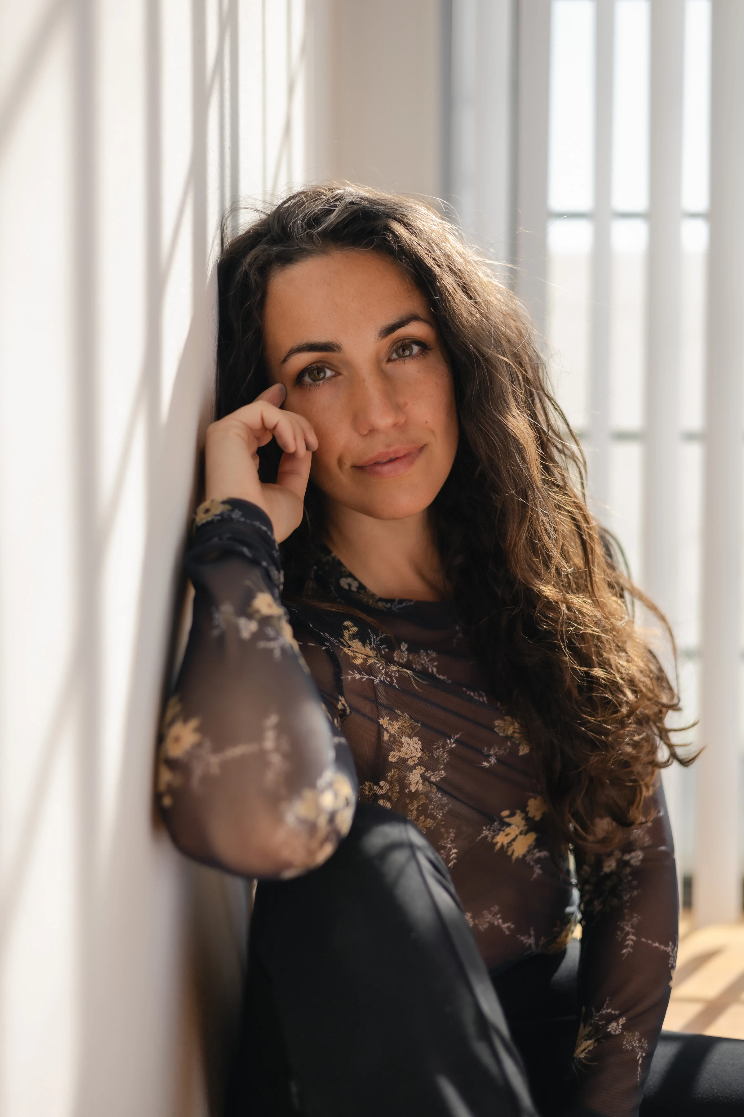 A woman with long wavy hair leans against a sunlit wall. She's wearing a sheer floral top, exuding a calm and contemplative mood.