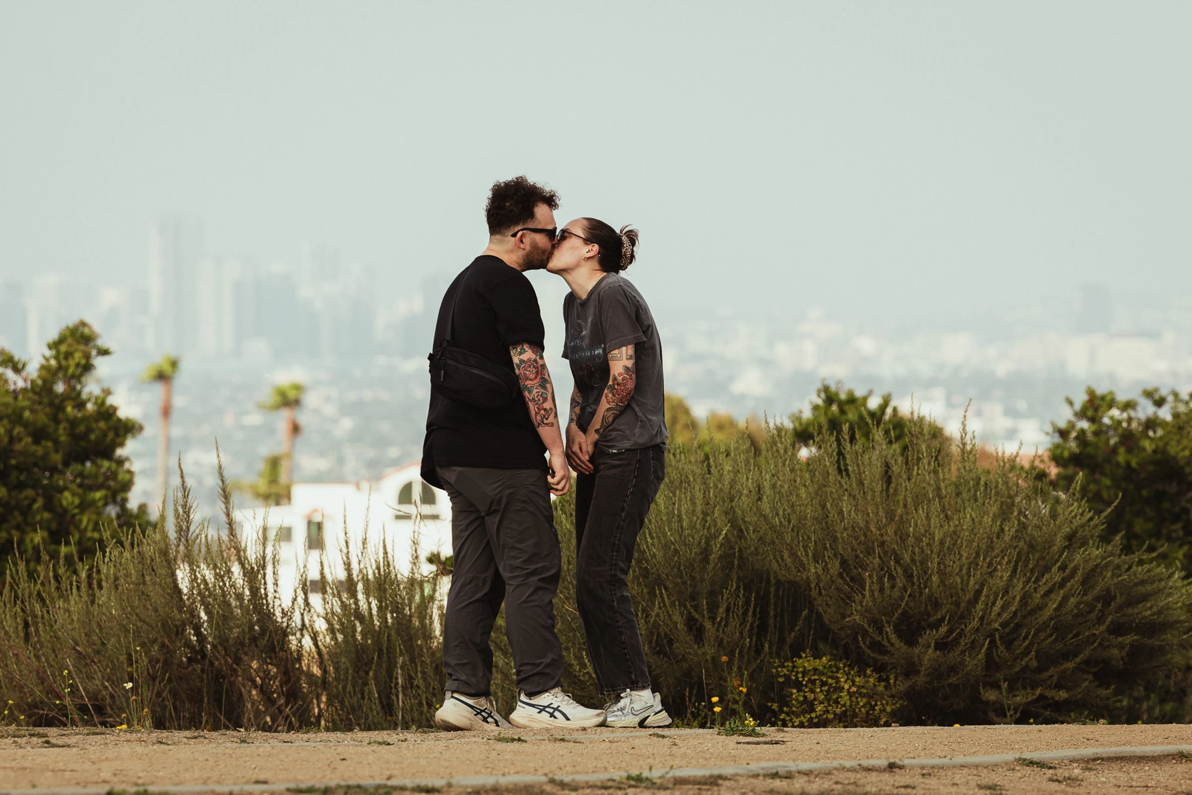 A couple with tattoos kisses outdoors, standing on a dirt path with greenery around. The city skyline is blurred in the background, conveying a peaceful, intimate moment.