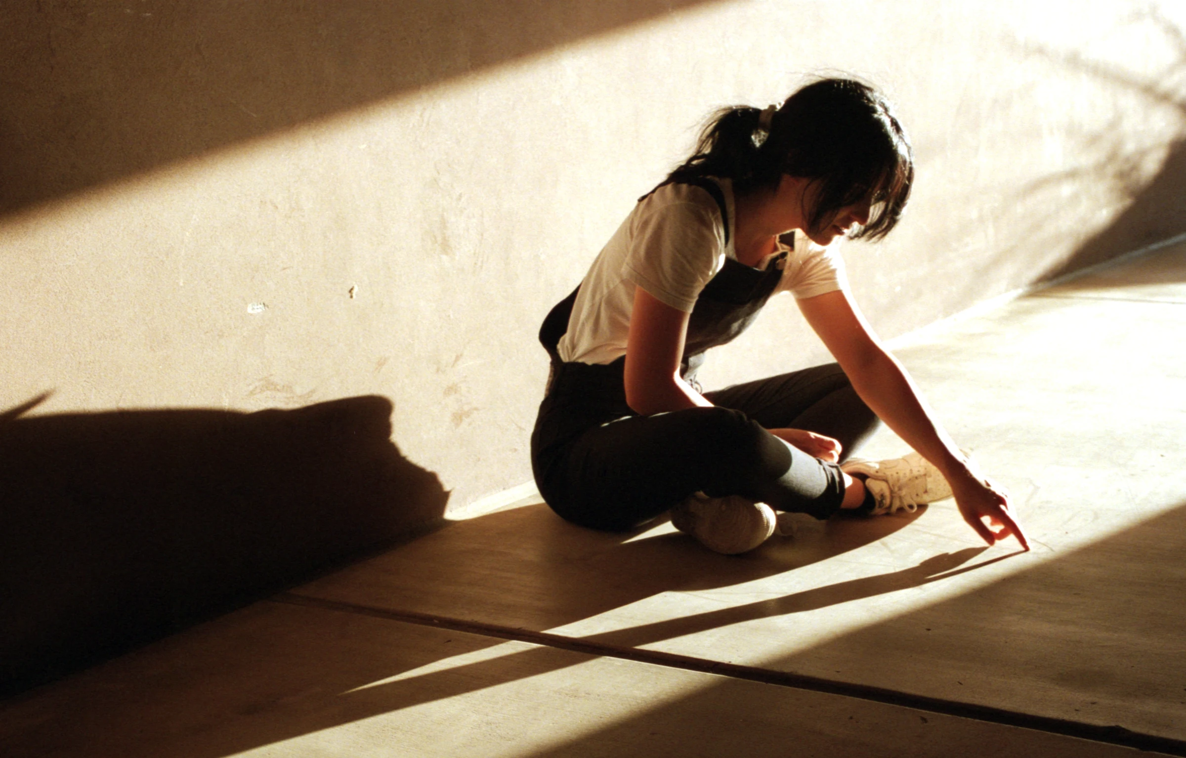 A woman sits on a sunlit floor, wearing a white shirt and dark overalls. She traces her finger along the ground, casting a thoughtful shadow.