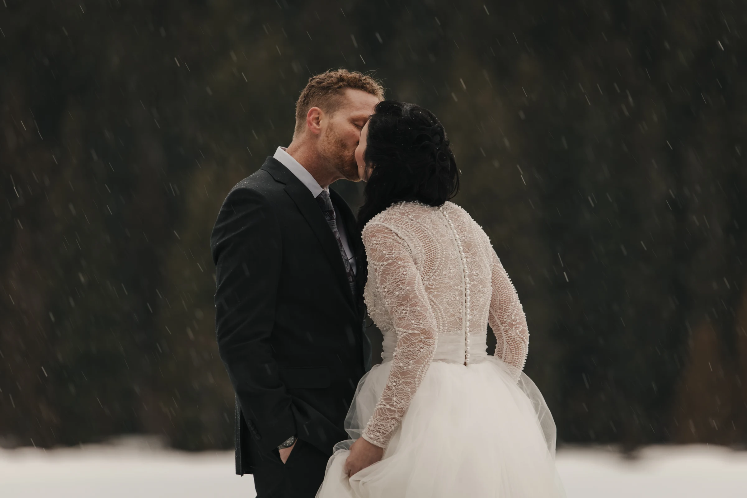 A bride and groom share a kiss in falling snow, surrounded by a blurred forest background. The bride wears a beaded dress, and the groom is in a dark suit. Romantic and serene.