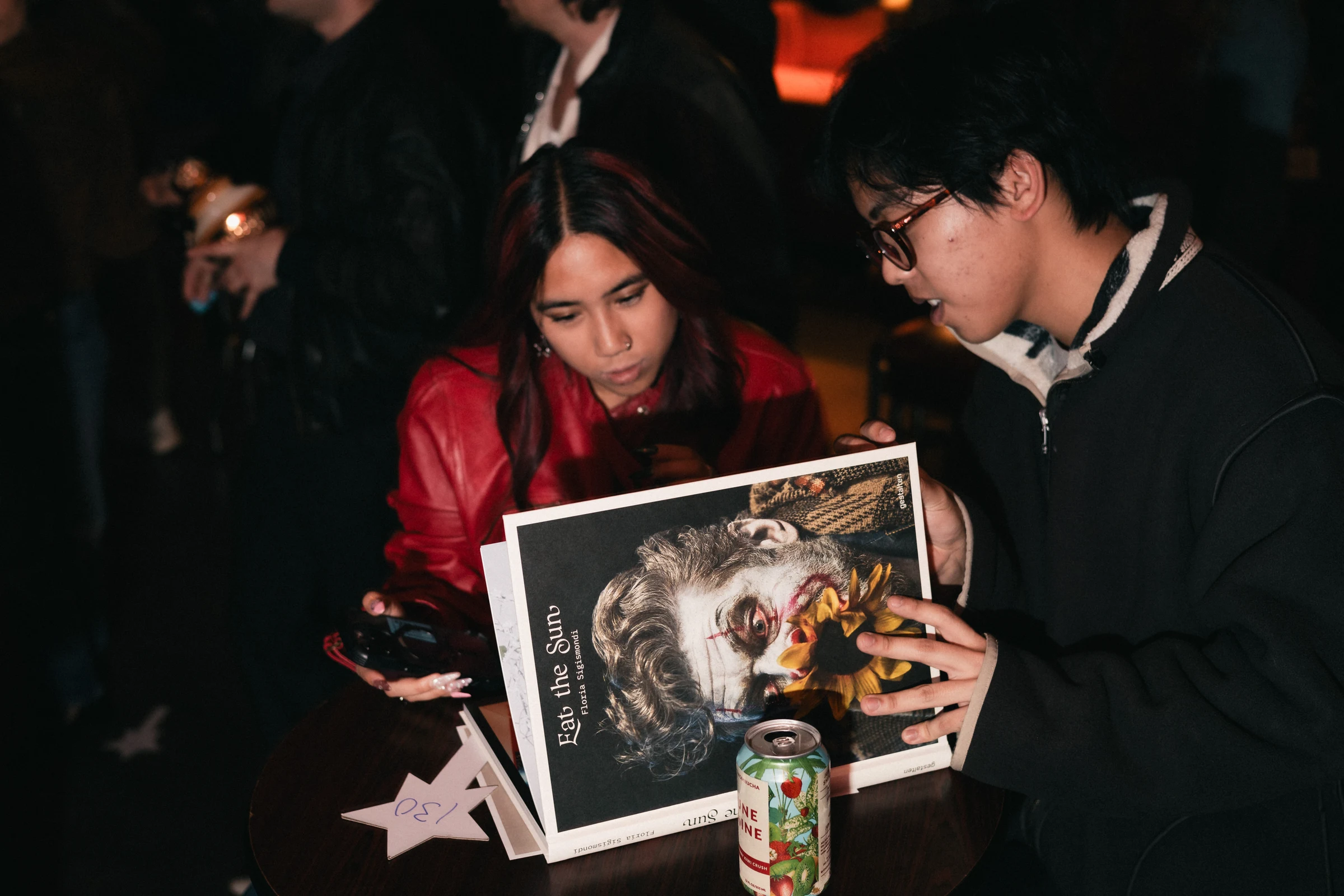 Two people are seated at a table, focused on a book with an artistic portrait on the cover. A can of drink and paper with numbers lie on the table, creating an engaged and thoughtful atmosphere.