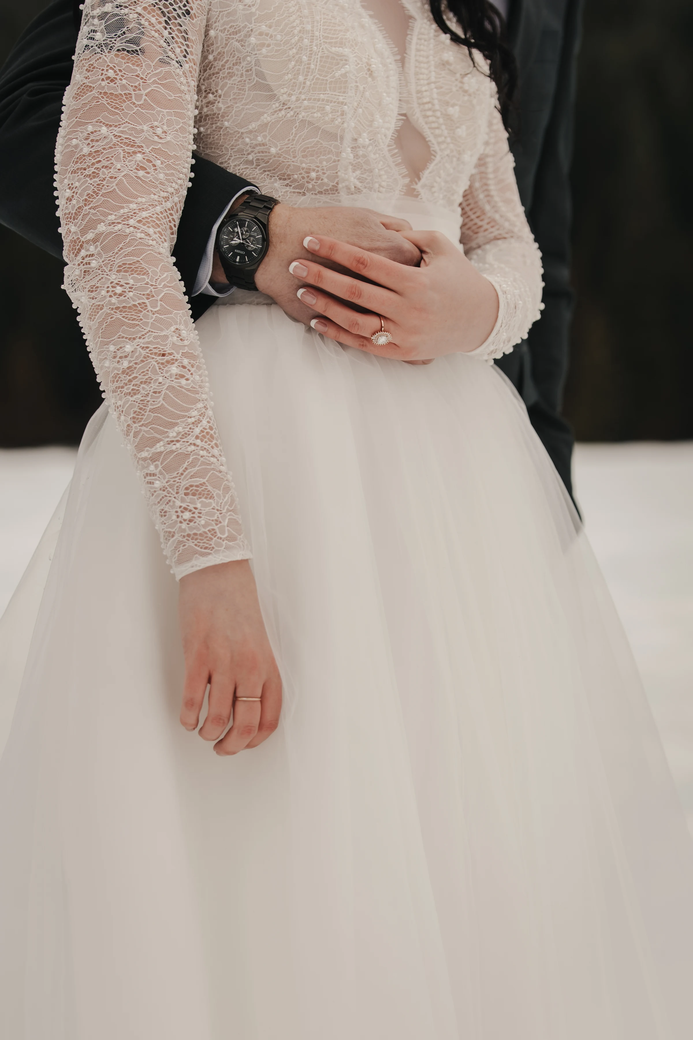 Close-up of a bride and groom. The bride is in a lace gown with a full skirt, her hands resting on the groom’s; he wears a watch. Romantic tone.