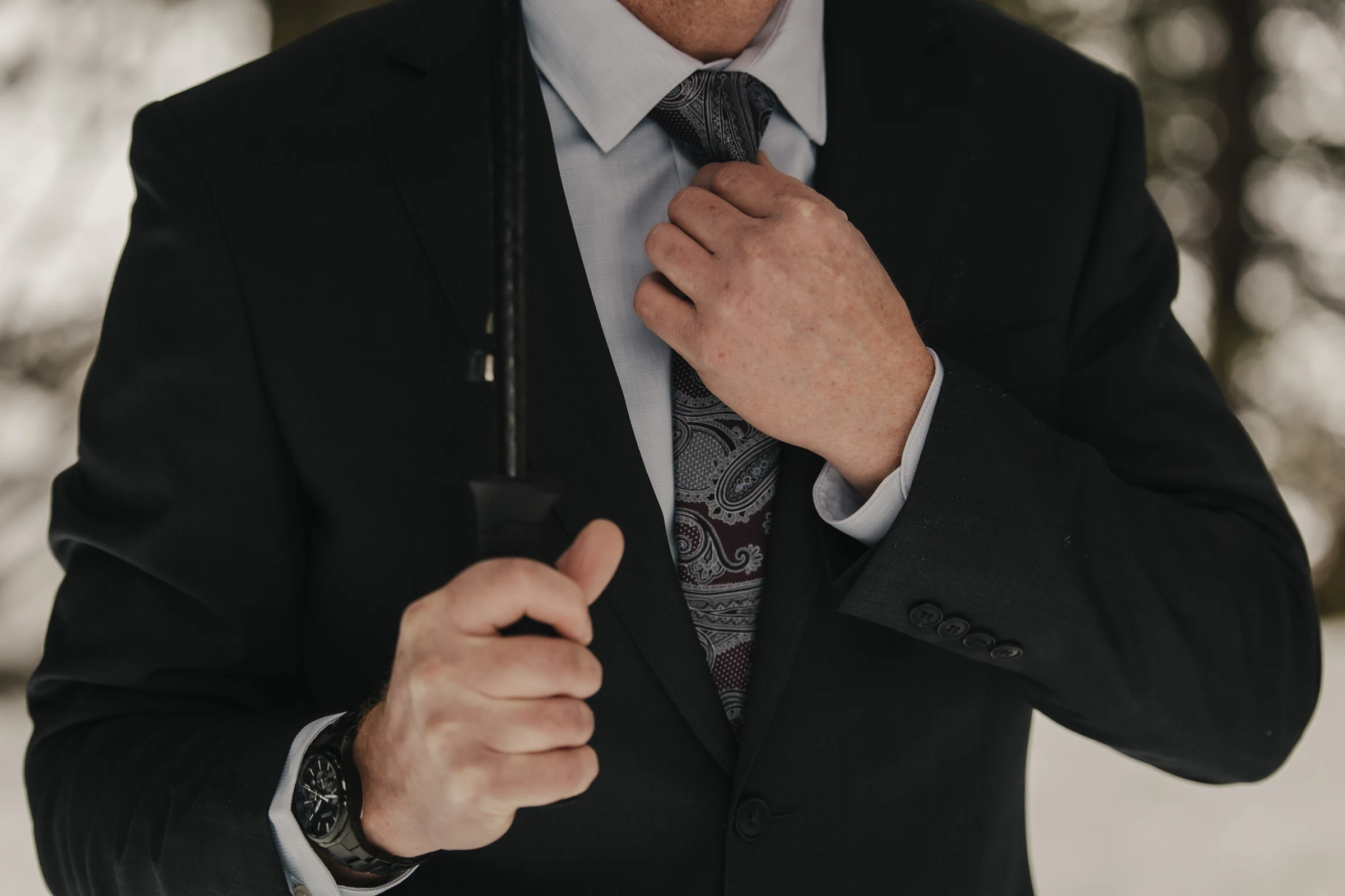 A man in a dark suit adjusts his paisley tie, holding a black umbrella. The setting appears outdoors with a blurred background, conveying a formal tone.