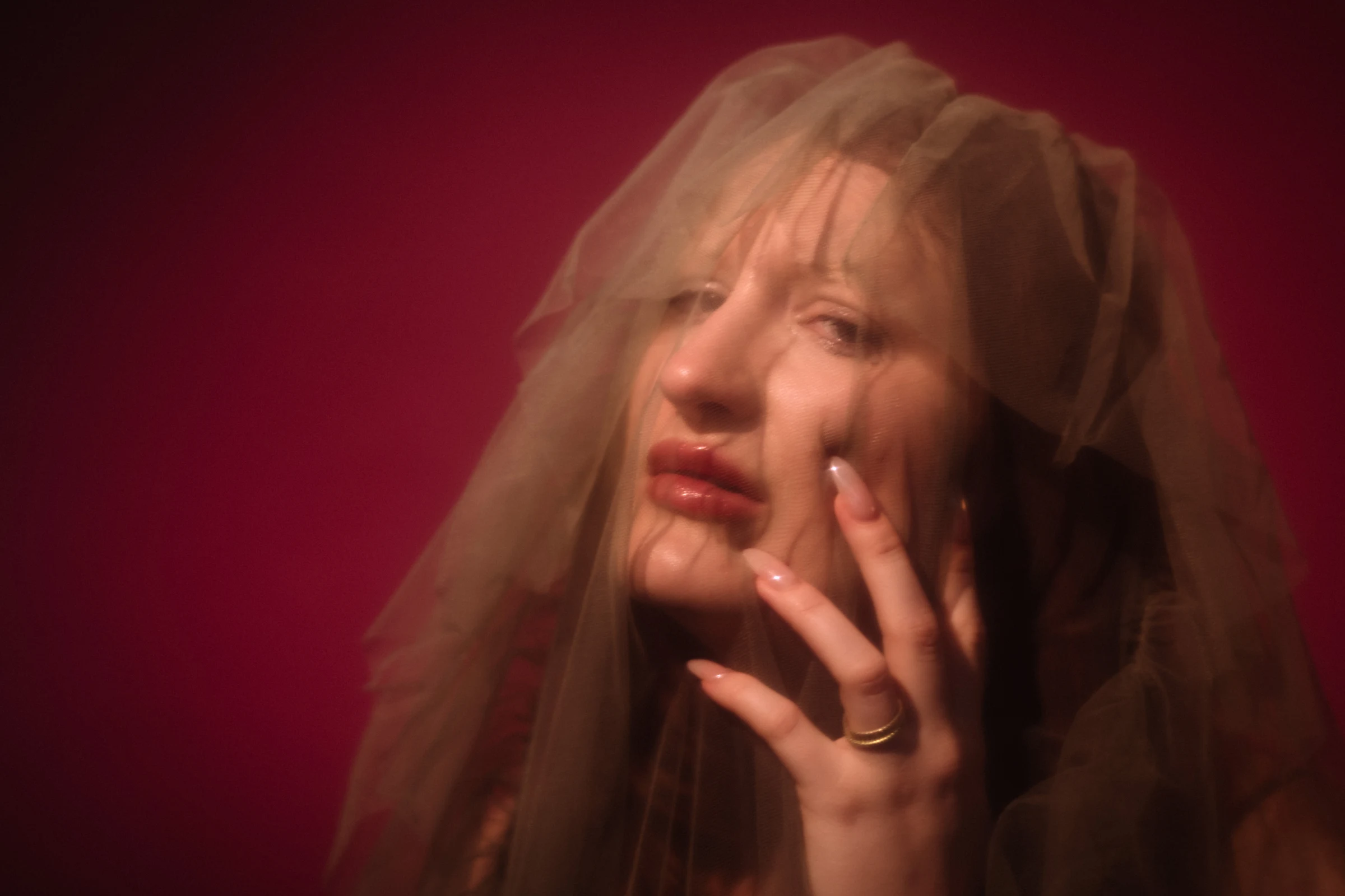 A woman with a sheer veil over her face, eyes closed and hand gently touching her cheek, exudes a dreamy, ethereal vibe against a deep pink background.