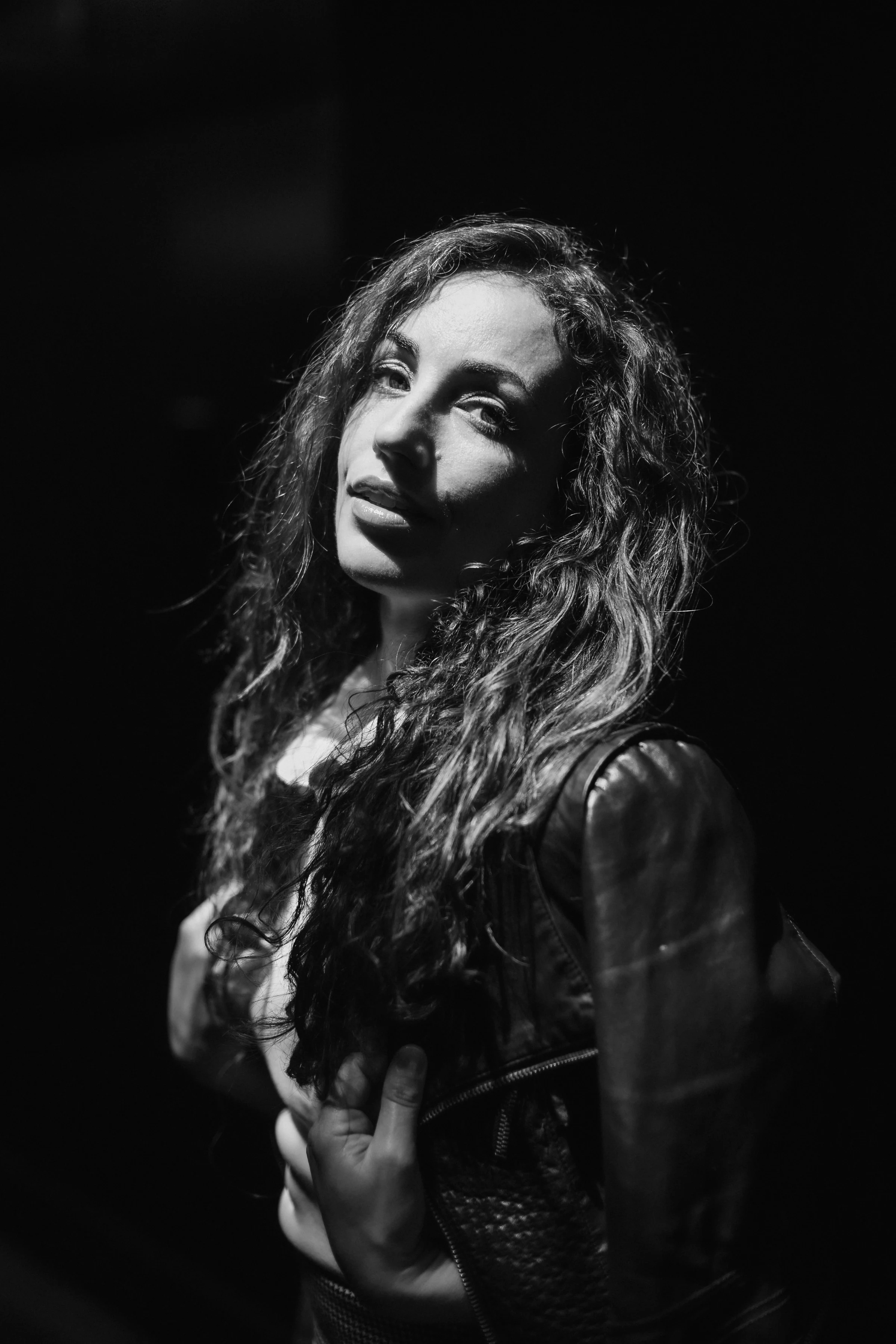 Black and white portrait of a woman with long, curly hair, illuminated by dramatic lighting. She gazes upward, wearing a leather jacket, conveying a mysterious and contemplative mood.