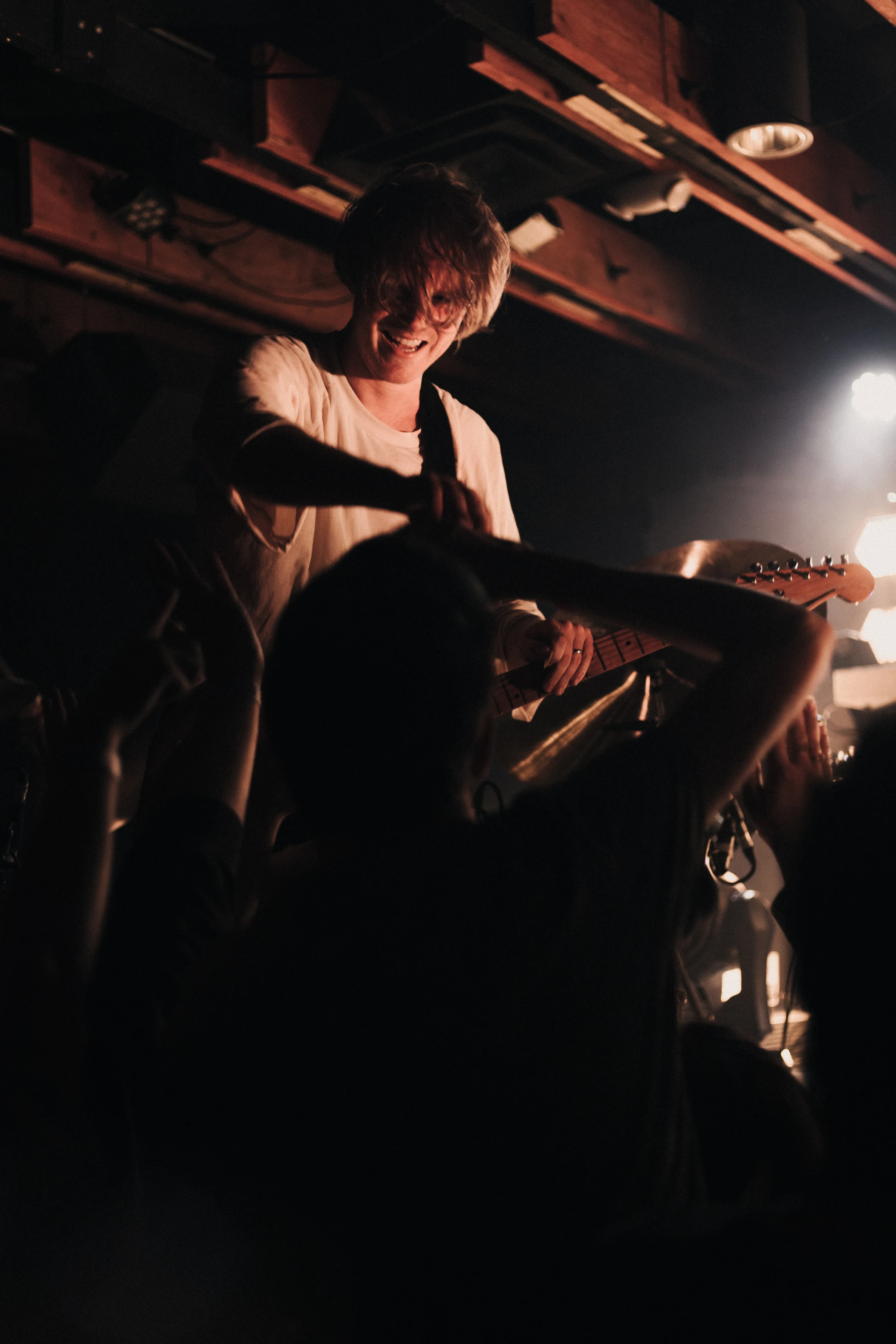 Musician with a guitar on stage, smiling and reaching towards an enthusiastic crowd in a dimly lit concert setting, creating an energetic and joyful atmosphere.