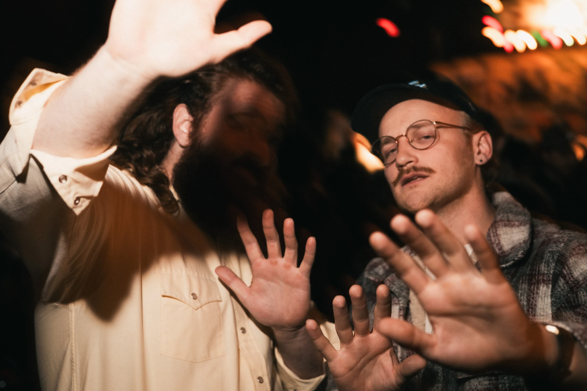 Two people with hands raised toward the camera in a dimly lit setting, partially obscuring their faces.