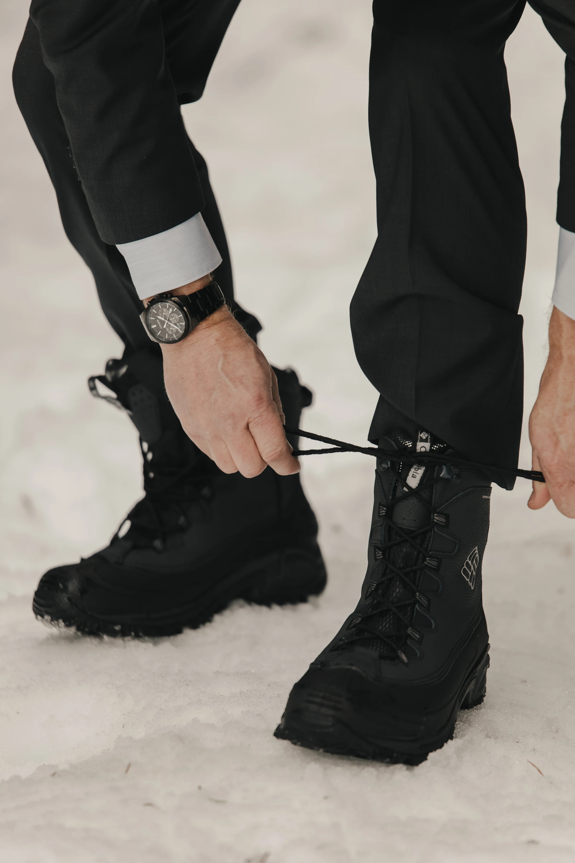A person in a suit ties the laces of black winter boots on a snowy surface. One wrist is adorned with a watch, conveying a blend of formal and outdoorsy style.