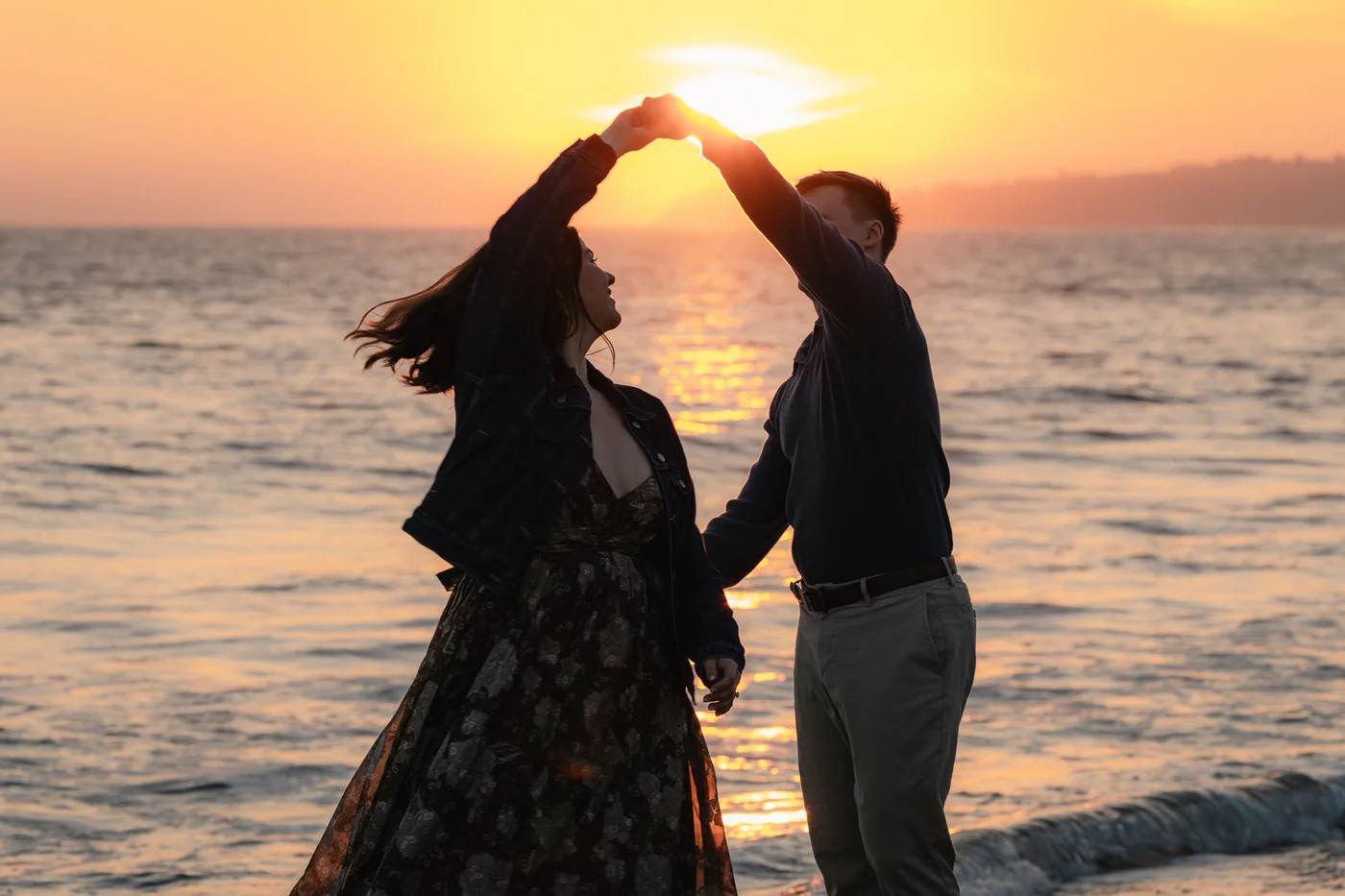 A couple dances joyfully by the ocean at sunset, silhouetted against a vibrant orange sky. The scene conveys romance and serenity.