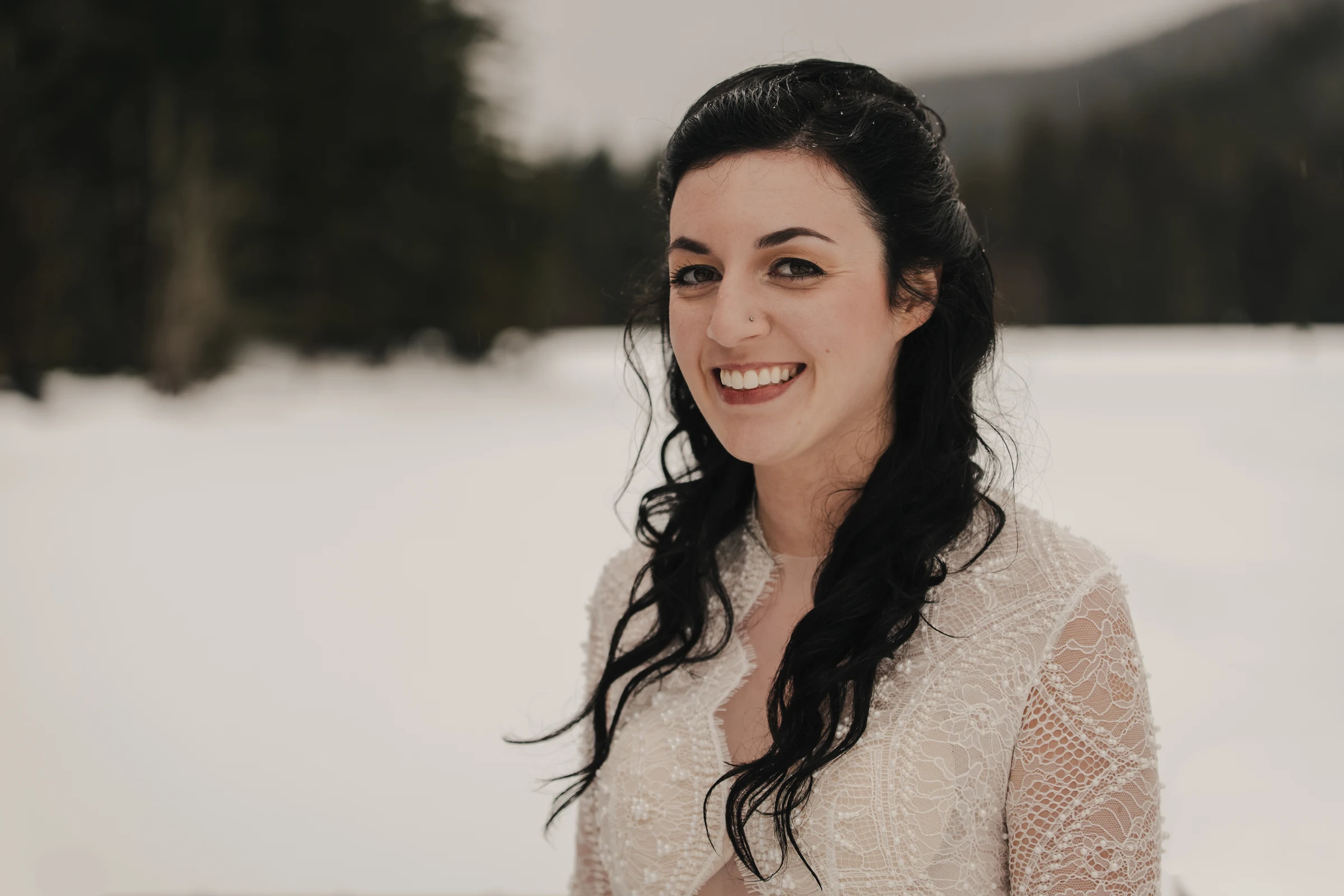 Smiling woman with long dark hair stands in a snowy landscape, wearing a lace dress. The scene is serene, with blurred trees in the background.