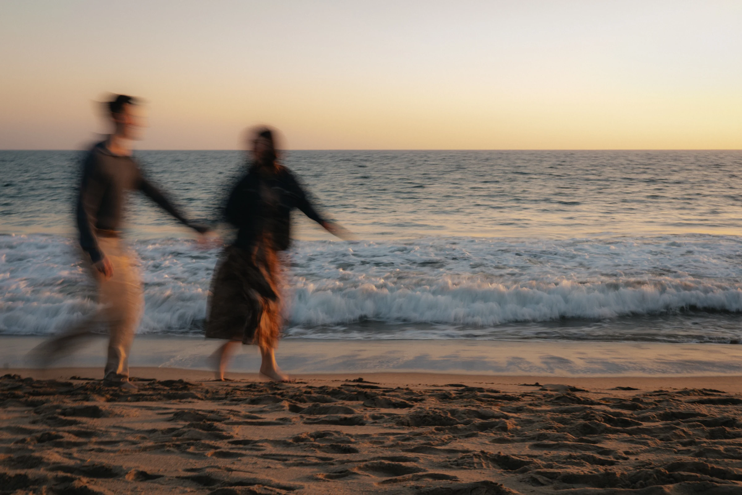 A blurred image of two people holding hands and walking along a sandy beach at sunset, with waves gently crashing in the background, creating a serene atmosphere.