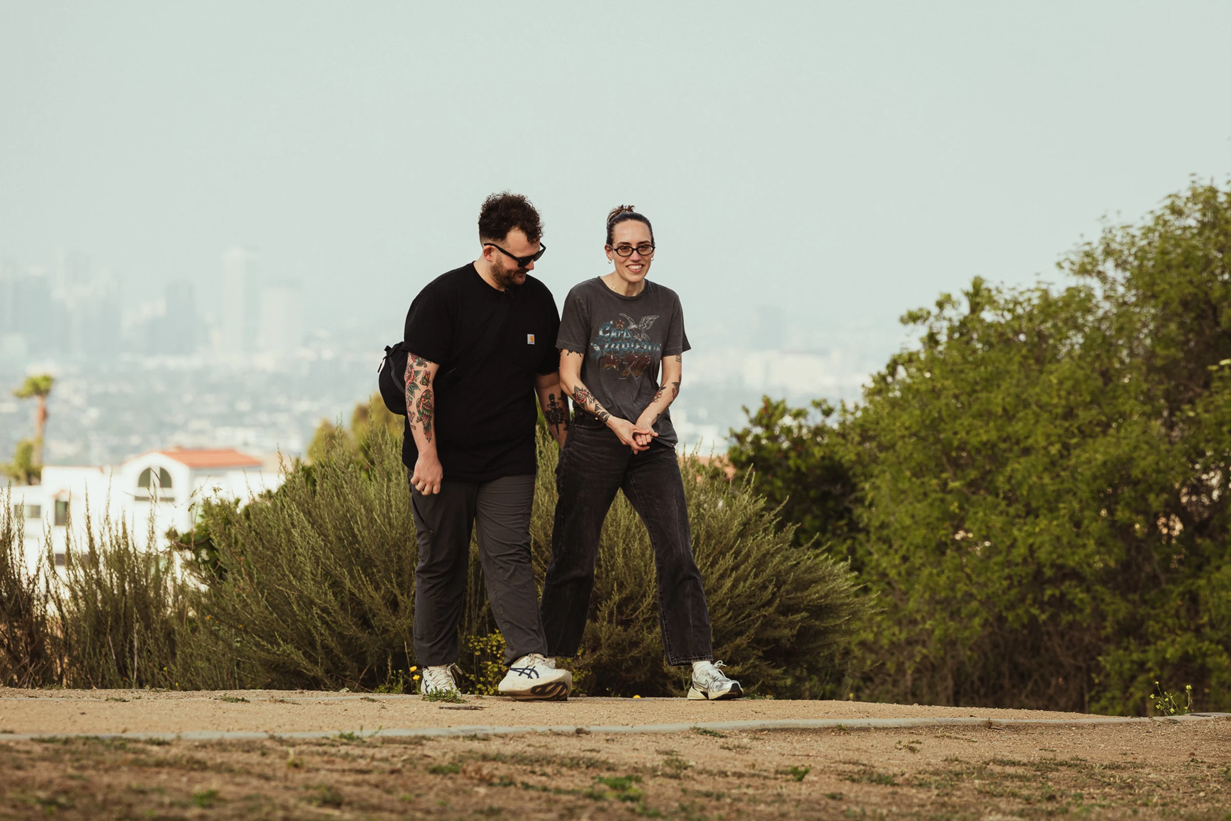 A man and a woman walk side by side on a dirt path surrounded by greenery, smiling and enjoying each other's company. The city skyline is blurred in the distance.