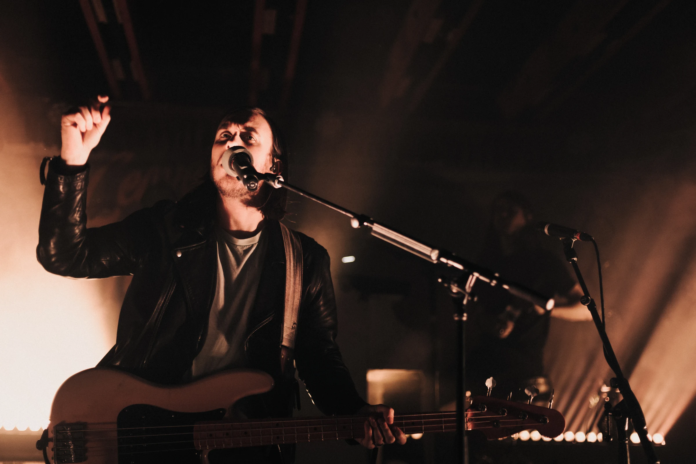 Musician in a dimly lit venue passionately sings into a microphone, holding a guitar. Warm stage lights create a moody, energetic atmosphere.