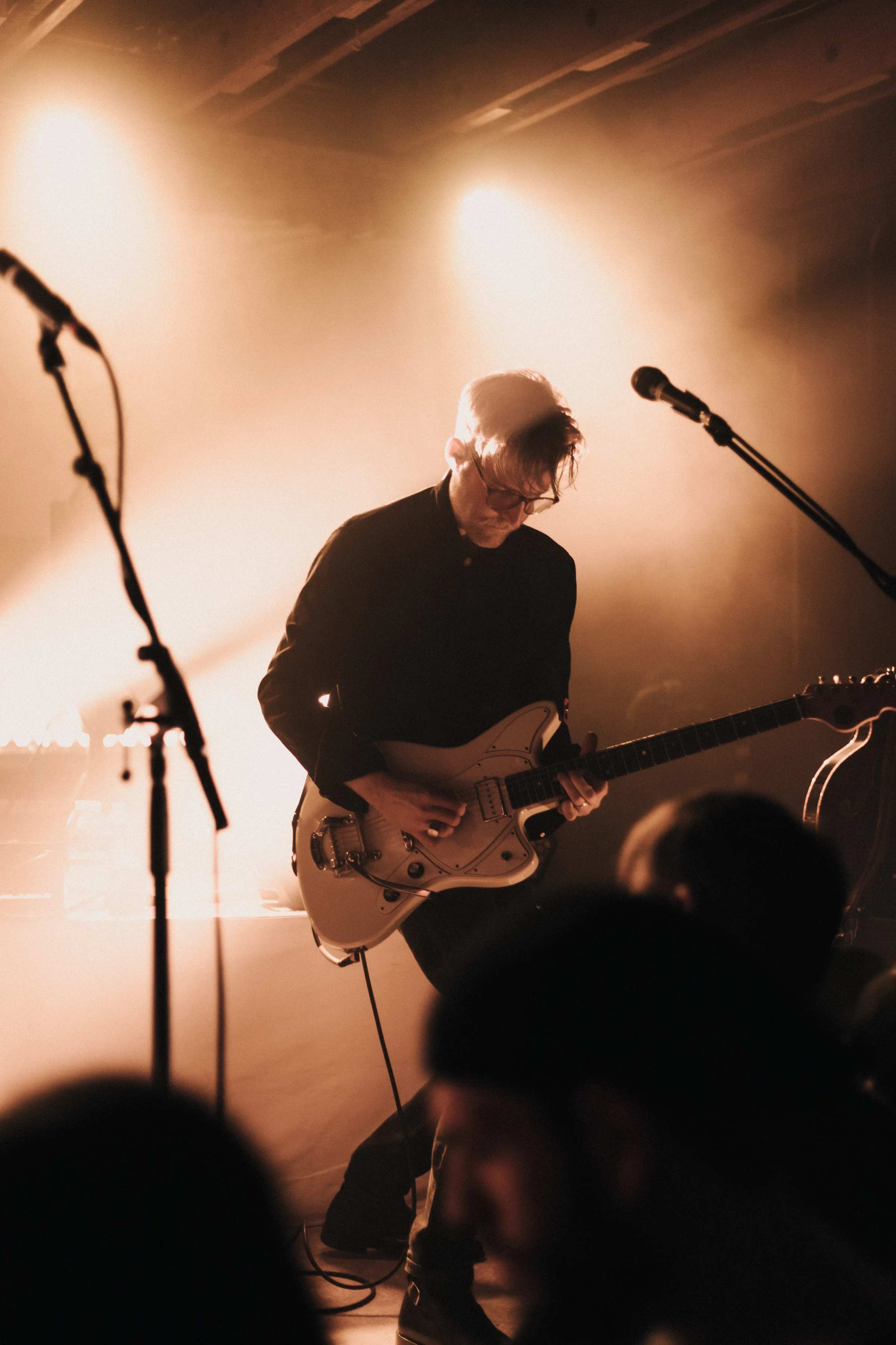 Musician plays electric guitar on stage under warm, atmospheric lighting. Silhouetted audience in the foreground creates an intimate concert vibe.