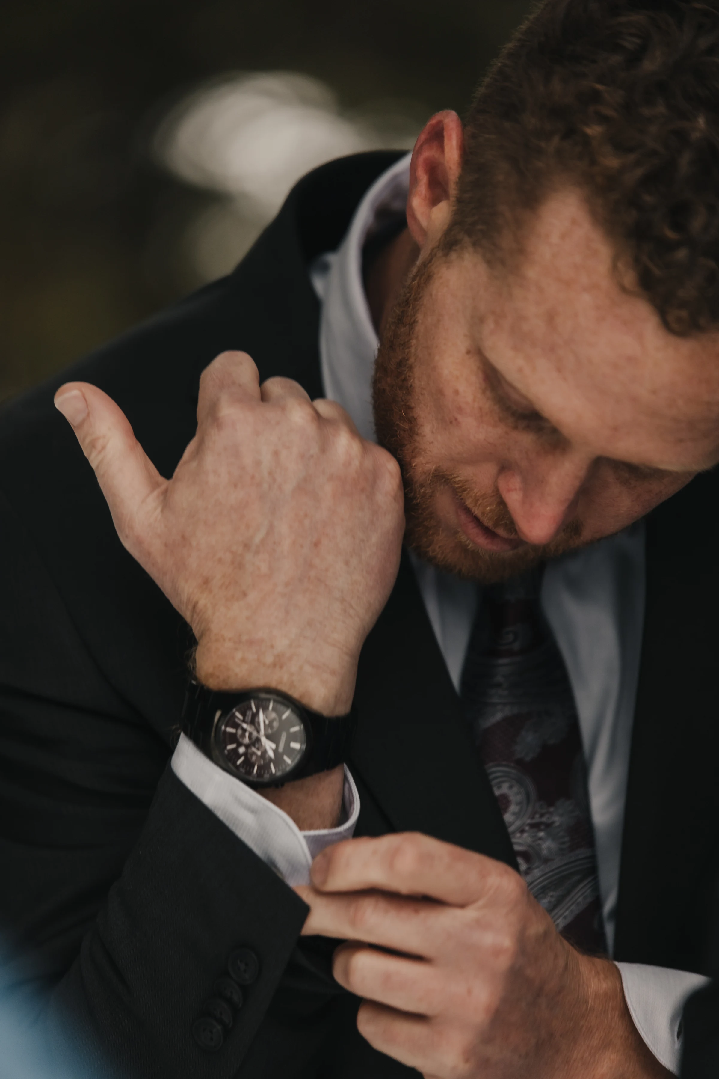 Man in a suit adjusting his shirt cuff, wearing a stylish watch. His expression is focused and introspective, suggesting a formal occasion.