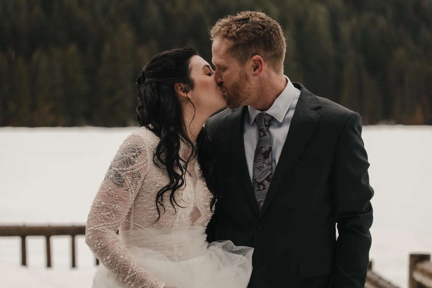 A couple kisses outdoors in a snowy setting, with a serene forest backdrop. The bride wears a lace wedding dress, and the groom is in a dark suit. Romantic and joyful atmosphere.