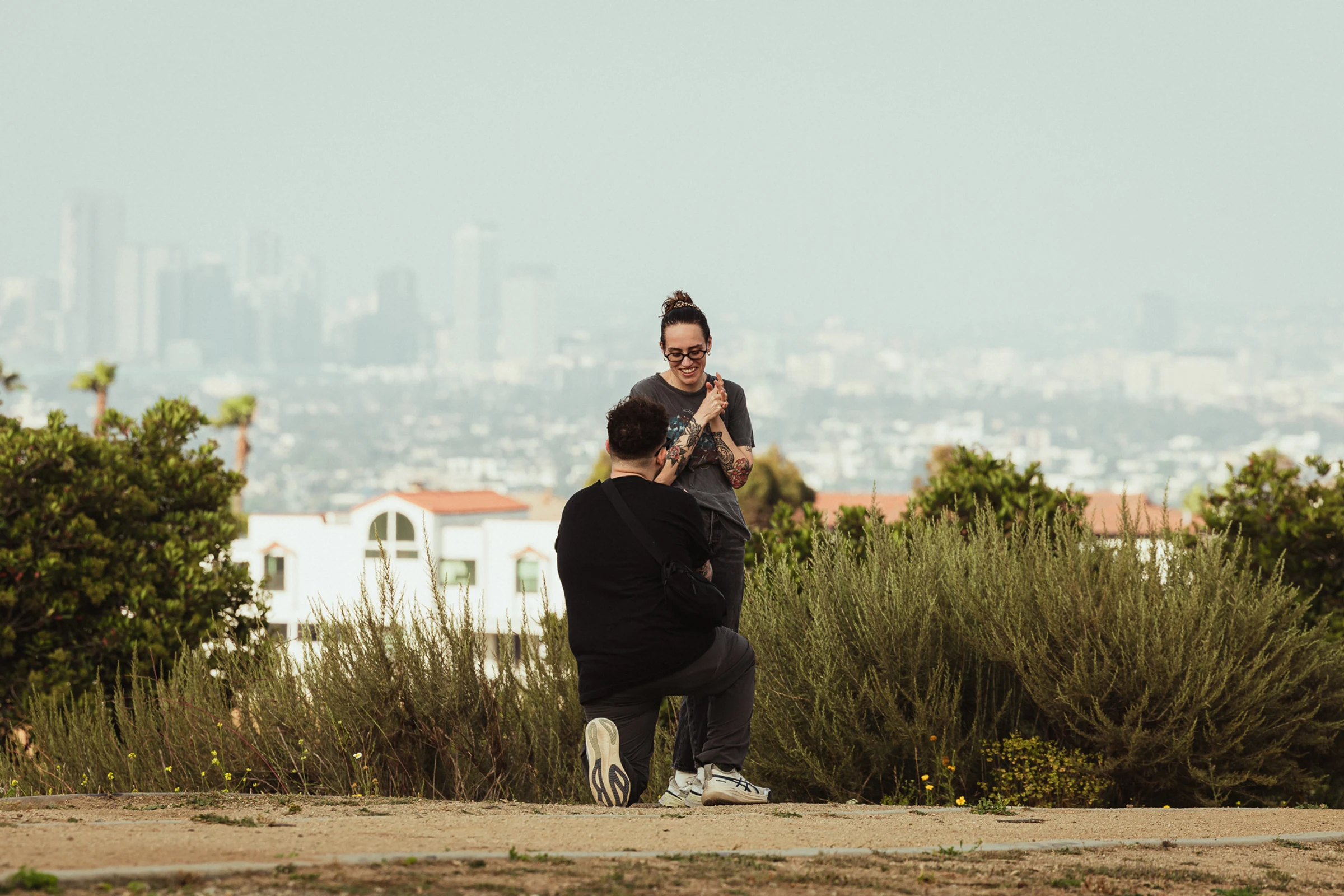 A person kneels to propose to a smiling partner in a park, with a cityscape in the background. The scene conveys love and excitement.